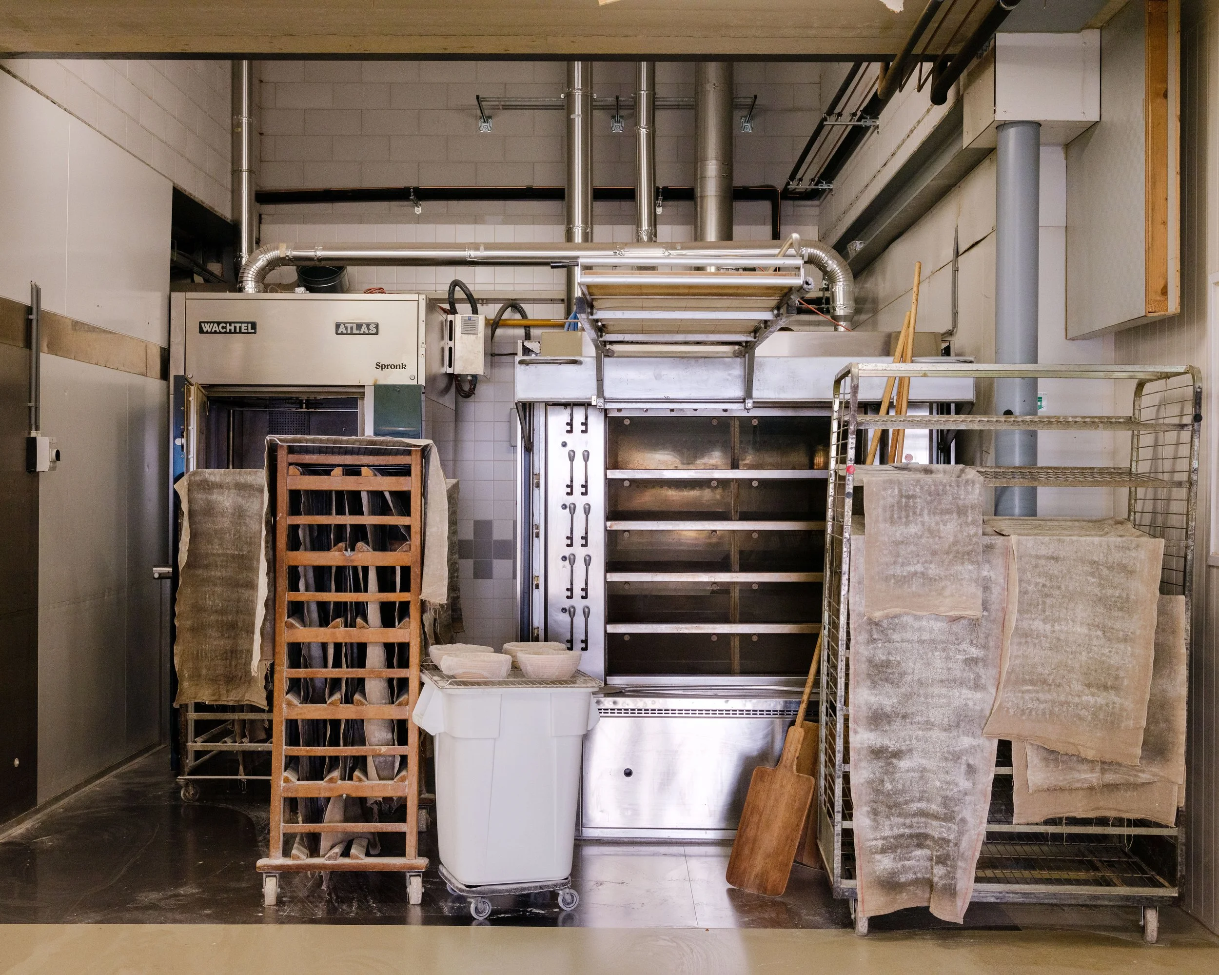 Industrial kitchen with stainless steel appliances, tools, and towels hanging on racks, including a large oven, shelving units, a rolling cart, and cleaning supplies.