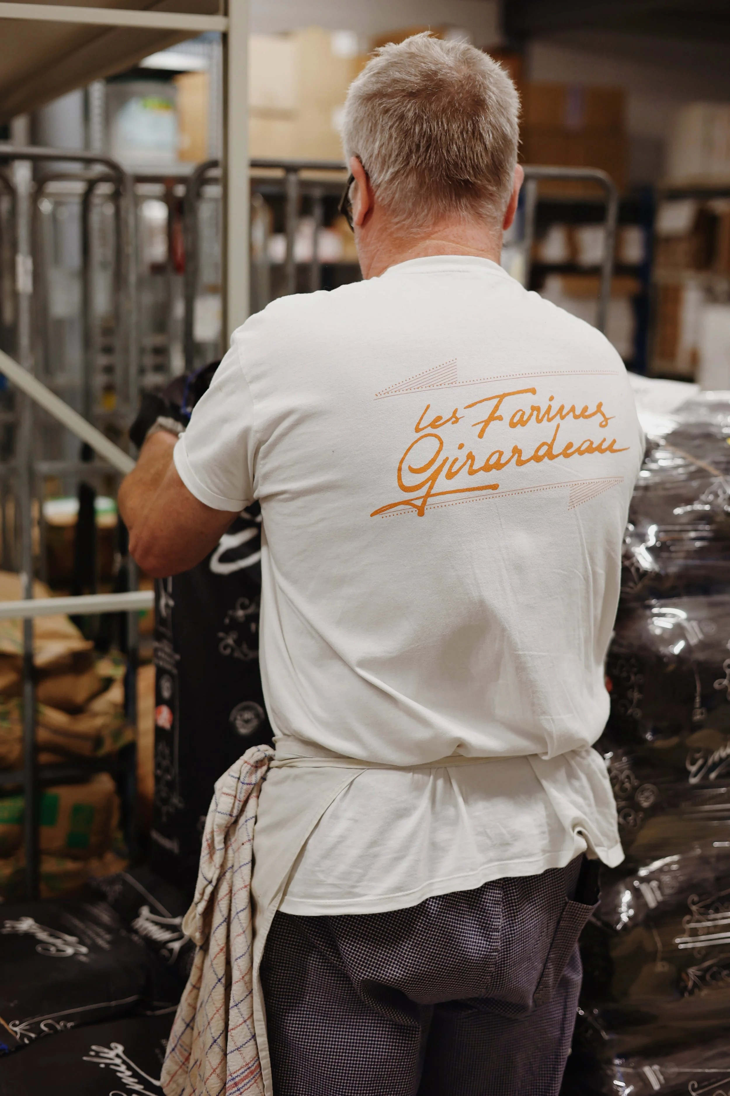 Back of a man wearing a white t-shirt with "Les Farires Girardeau" printed on it, working in a storage room or warehouse surrounded by boxes and packaging materials.