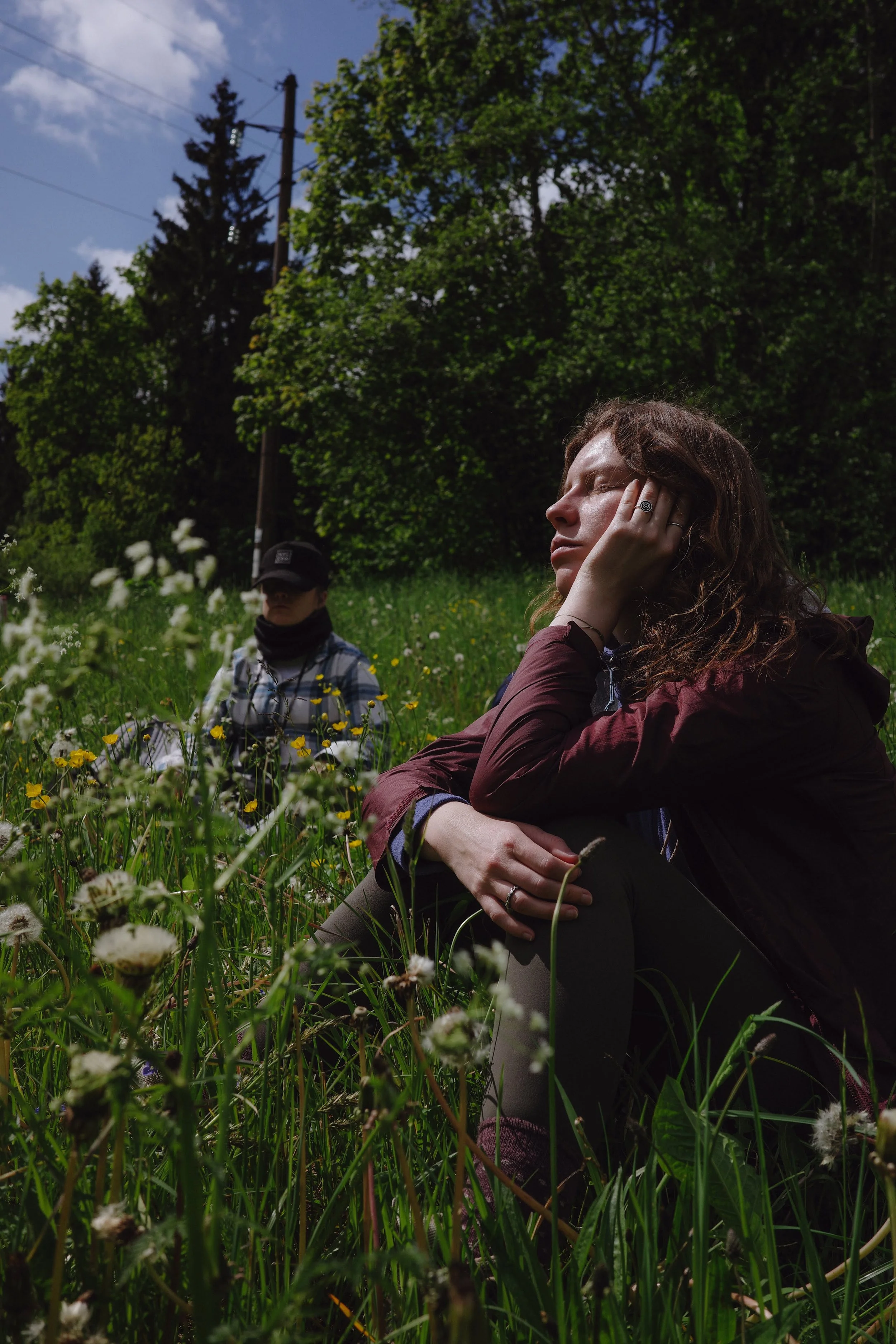 Two women sitting on grassy field surrounded by wildflowers, one with hand on face, eyes closed, trees and utility pole in background on a sunny day.