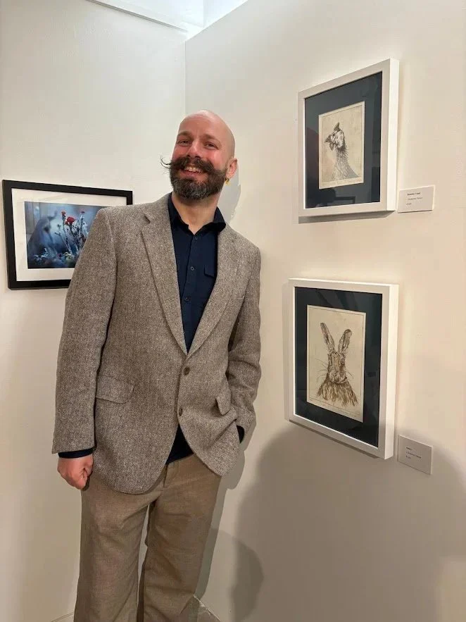 A man with a beard and a bald head, dressed in a tweed blazer and beige pants, stands in an art gallery beside framed artwork, smiling at the camera.
