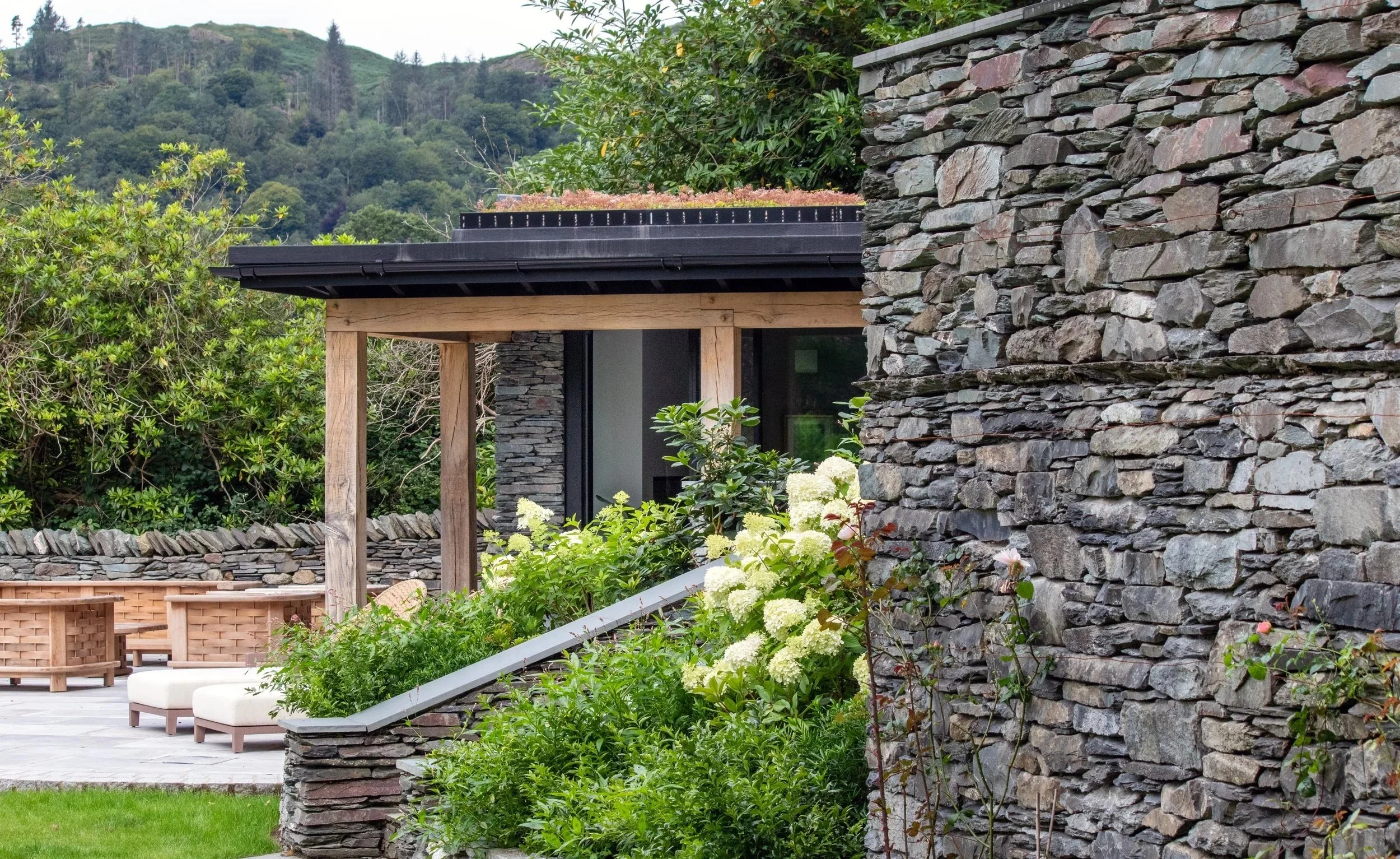 A modern outdoor patio area with stone walls, wooden structures, and lush green plants surrounding a stone pathway and seating area.
