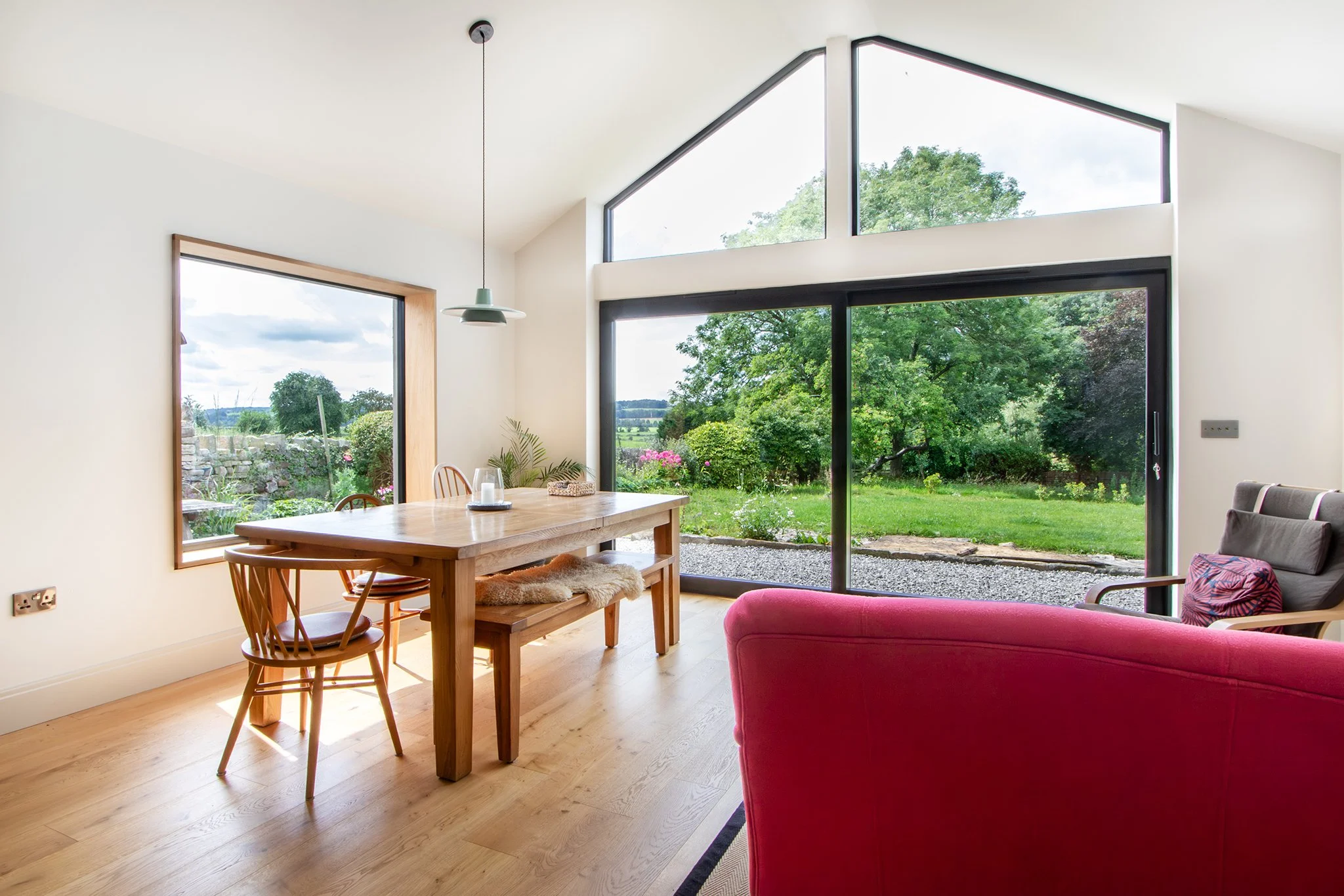 Bright living room with large floor-to-ceiling windows overlooking a green garden, wooden dining table with chairs, red armchair, and modern decor.