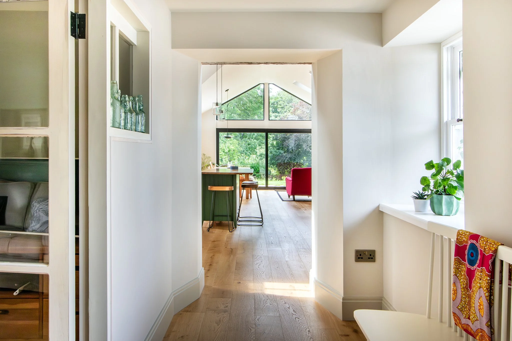 Interior view of a bright, modern living space with large windows, wooden flooring, and minimalist decor including a green kitchen island and a red armchair.