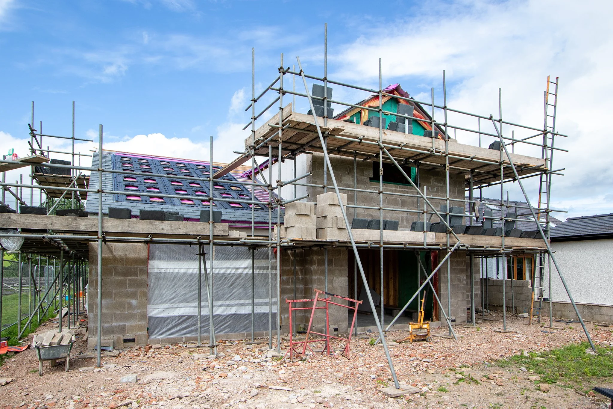 Under construction house with scaffolding, partial roof and brick walls, on a gravel lot with a blue sky and clouds.