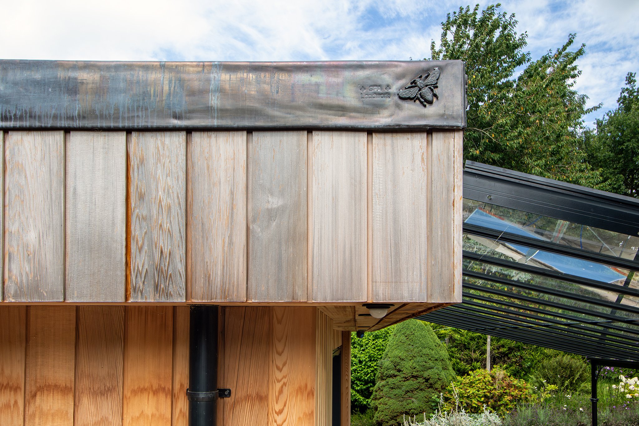 Close-up of a modern wooden building with a metal roof corner, some trees, and plants in the background.