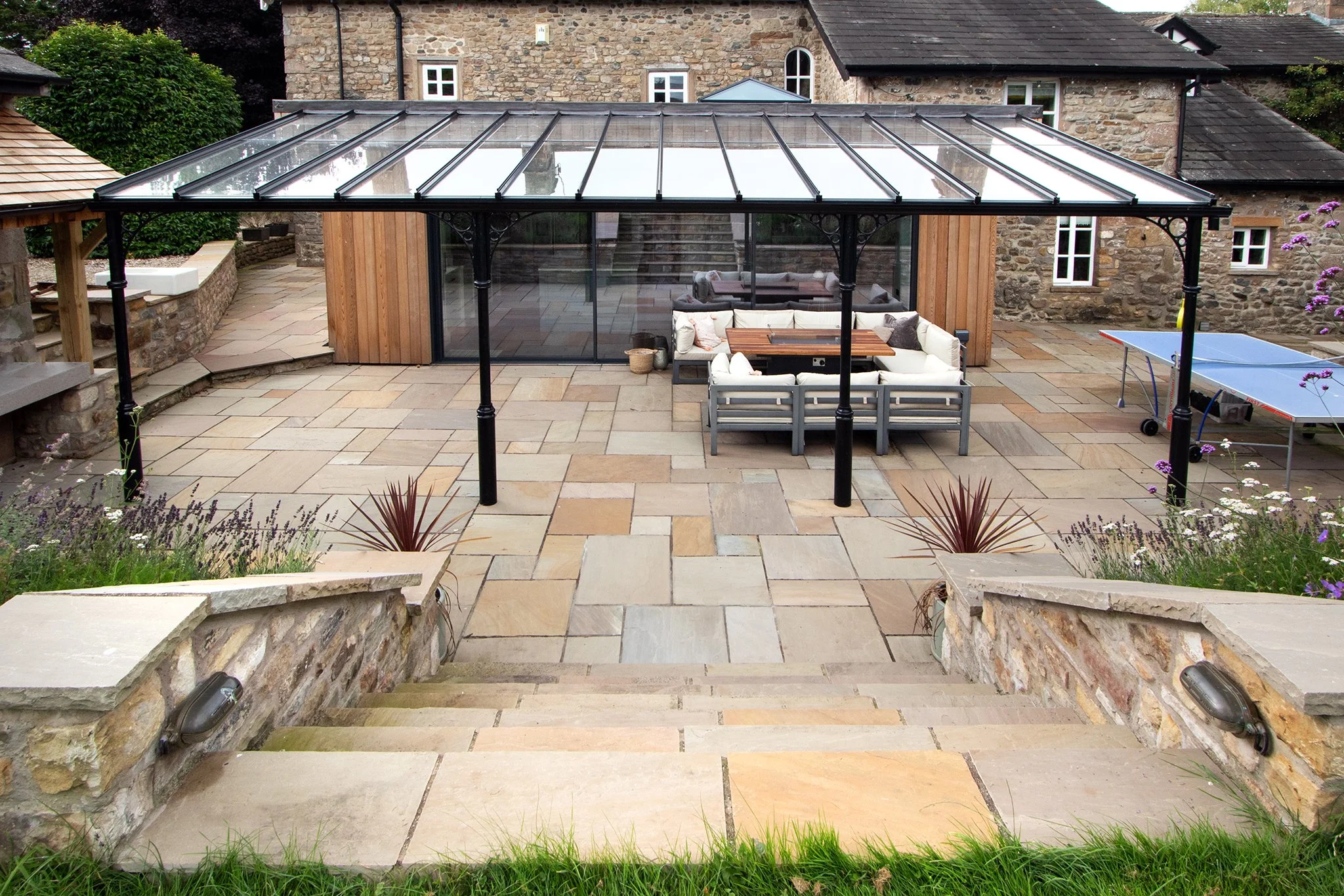 Stone steps leading down to a paved patio with a black metal framed glass roof, outdoor seating area with cushioned furniture, and a ping pong table. Stone and brick buildings are in the background.