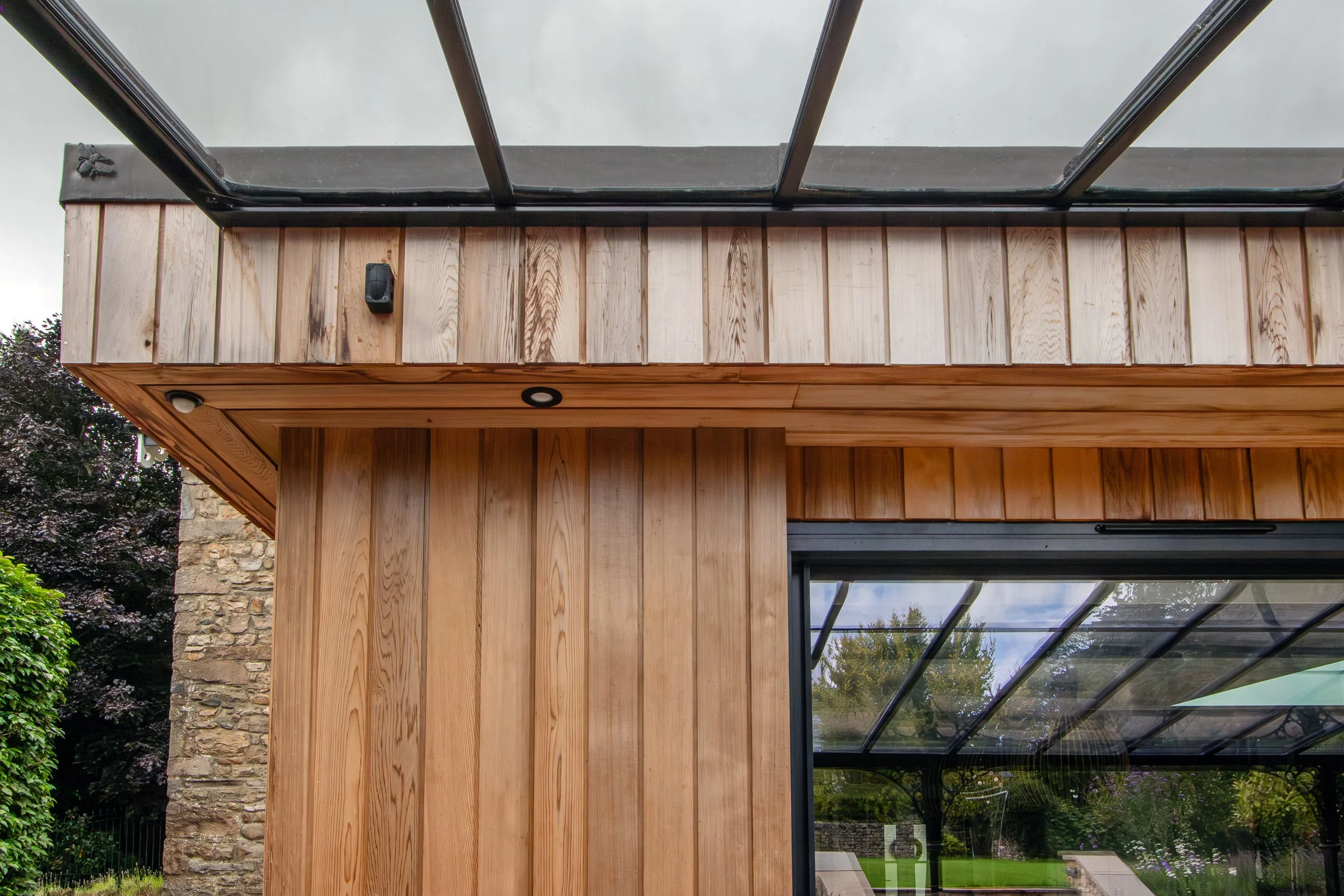 Close-up view of a building corner with wooden siding and a glass roof, overlooking a landscaped garden.