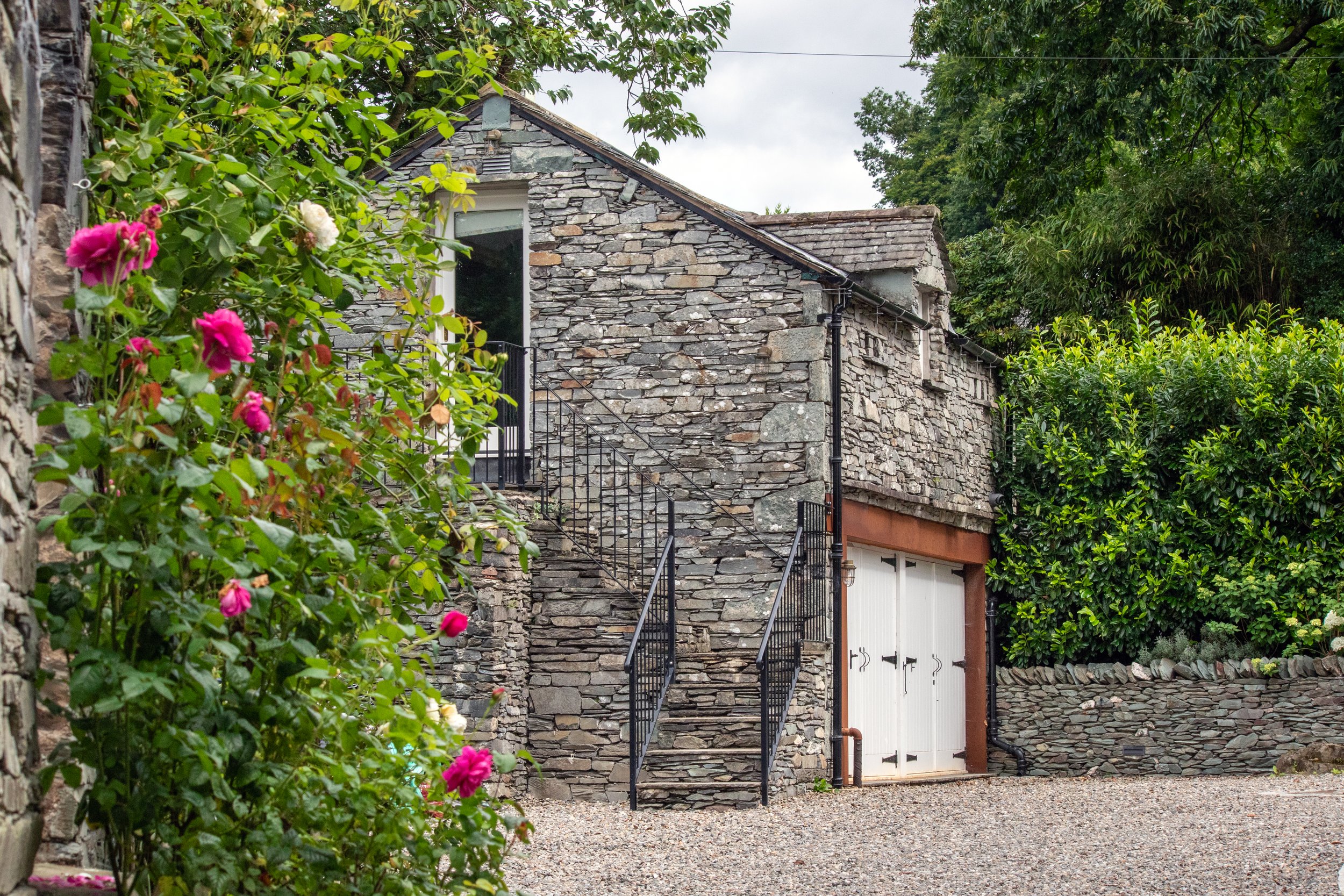 A stone house with a staircase, a white garage door, greenery, and flowering plants