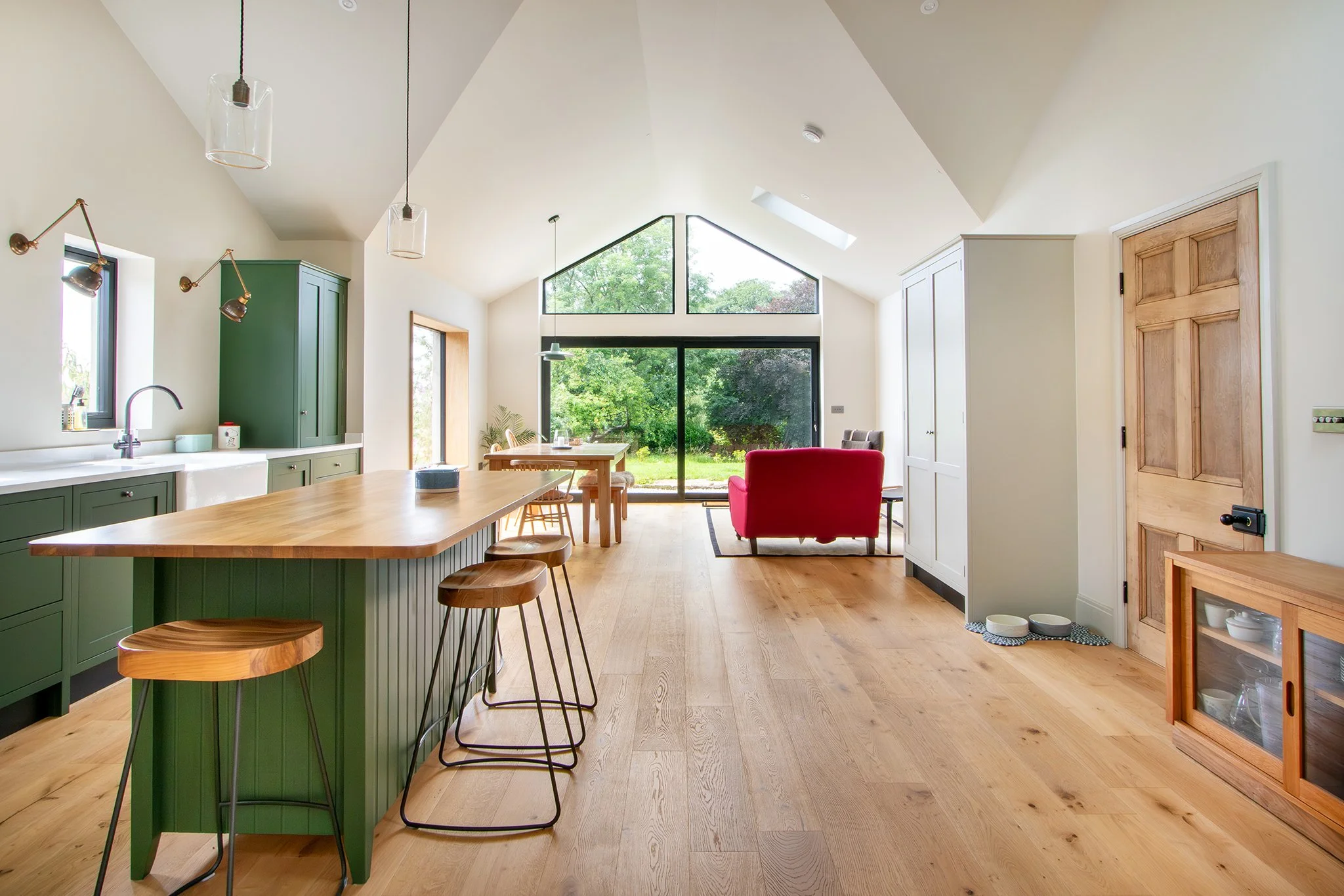 Open-concept kitchen and living area with green cabinets, wooden island, dining table, red loveseat, large window, and hardwood floors.