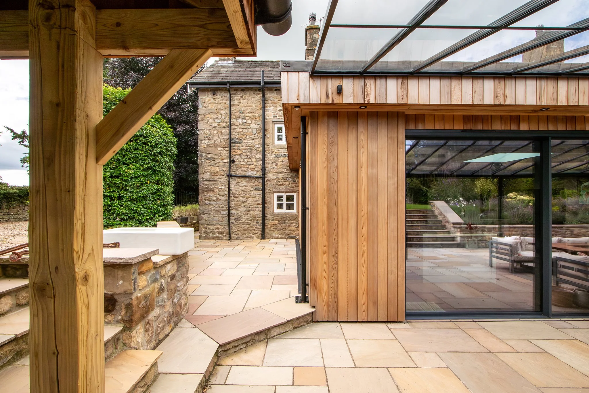 Outdoor patio area with stone and wooden structures, large glass sliding door, and steps leading up to another level, surrounded by greenery.