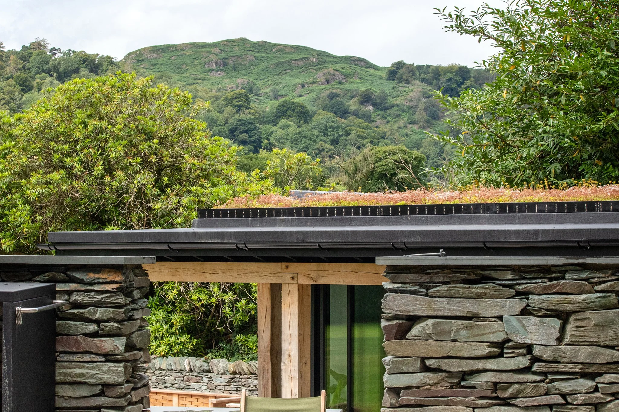 Close-up of a modern building with stone walls, black metal roof, glass door, and a wooden support beam, with lush green trees and hillside in the background.