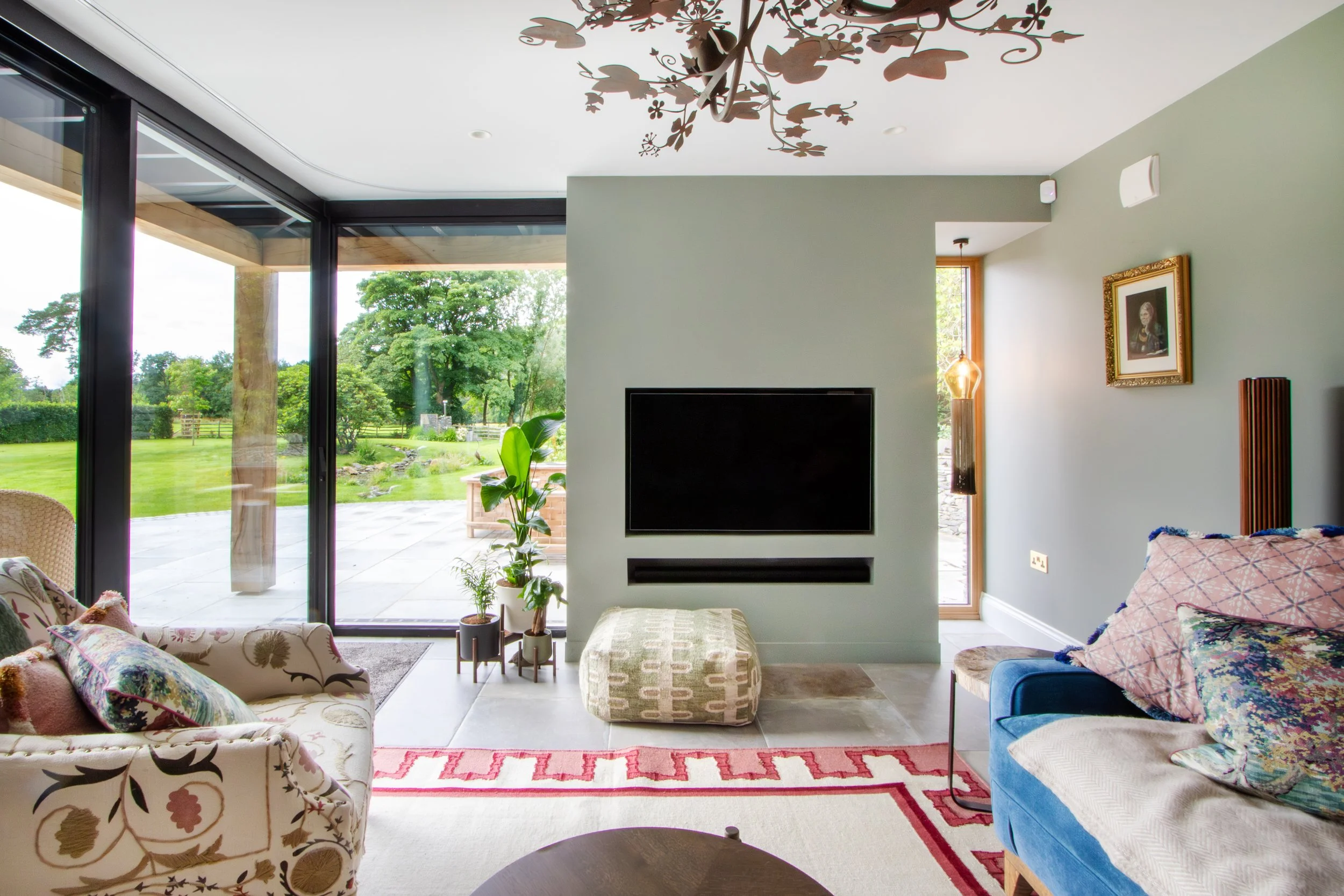 Living room with large glass sliding doors opening to a green garden, wall-mounted television, patterned sofas, and various potted plants.
