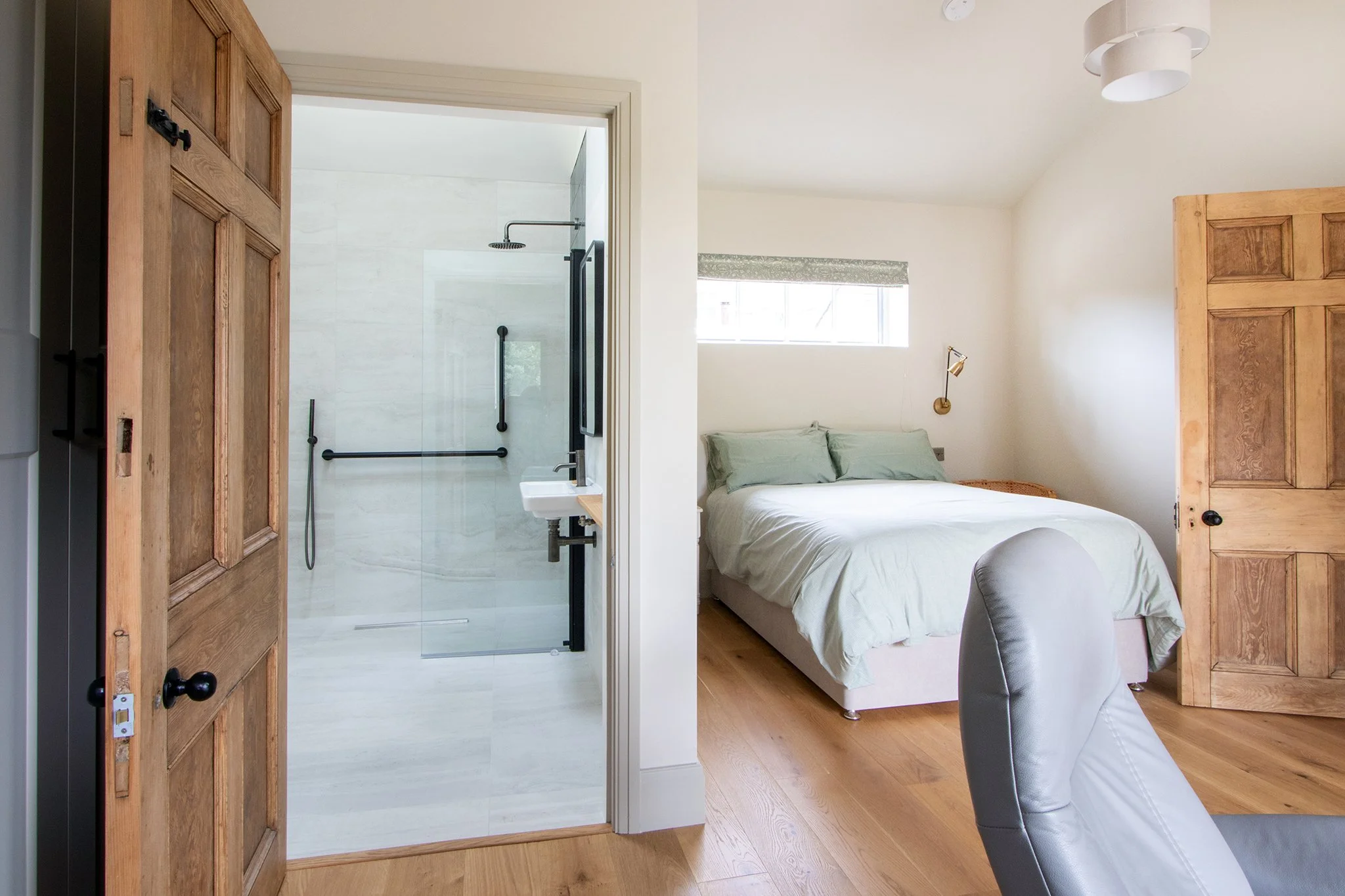 A bedroom with a white bed, wooden floors, and a wall-mounted lamp; adjacent bathroom with a walk-in shower with black fixtures, visible through a glass door.