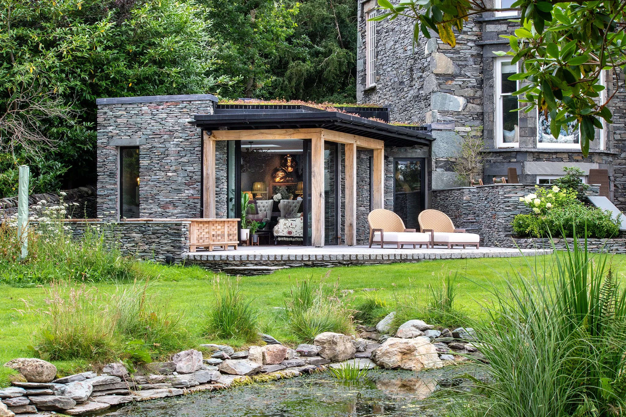 A modern house with a stone exterior, large windows, and a backyard patio with outdoor seating, overlooking a pond with rocks and greenery.