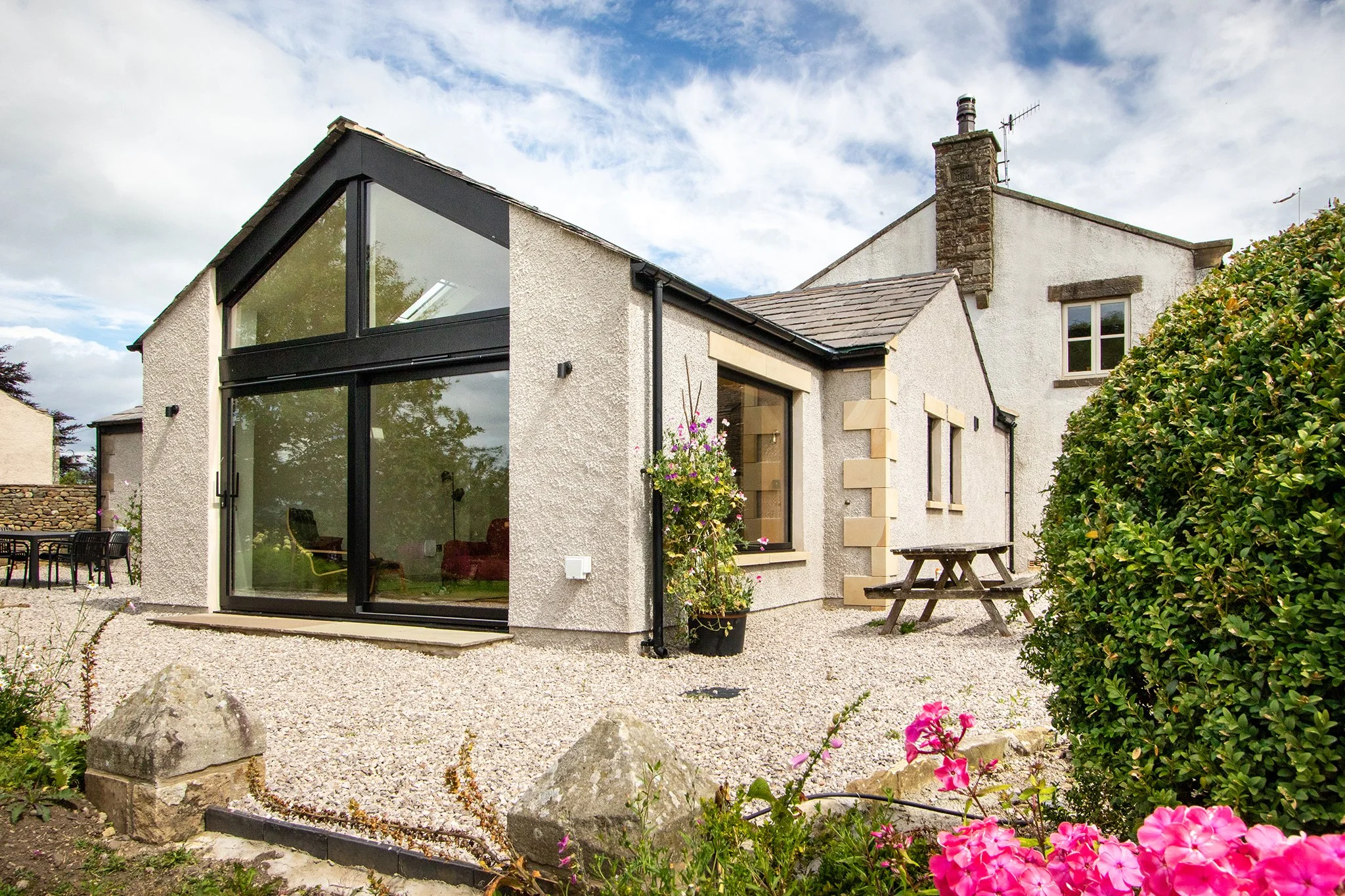 Exterior of a house with a modern glass extension, a gravel yard, a picnic table, and blooming pink flowers in the foreground.