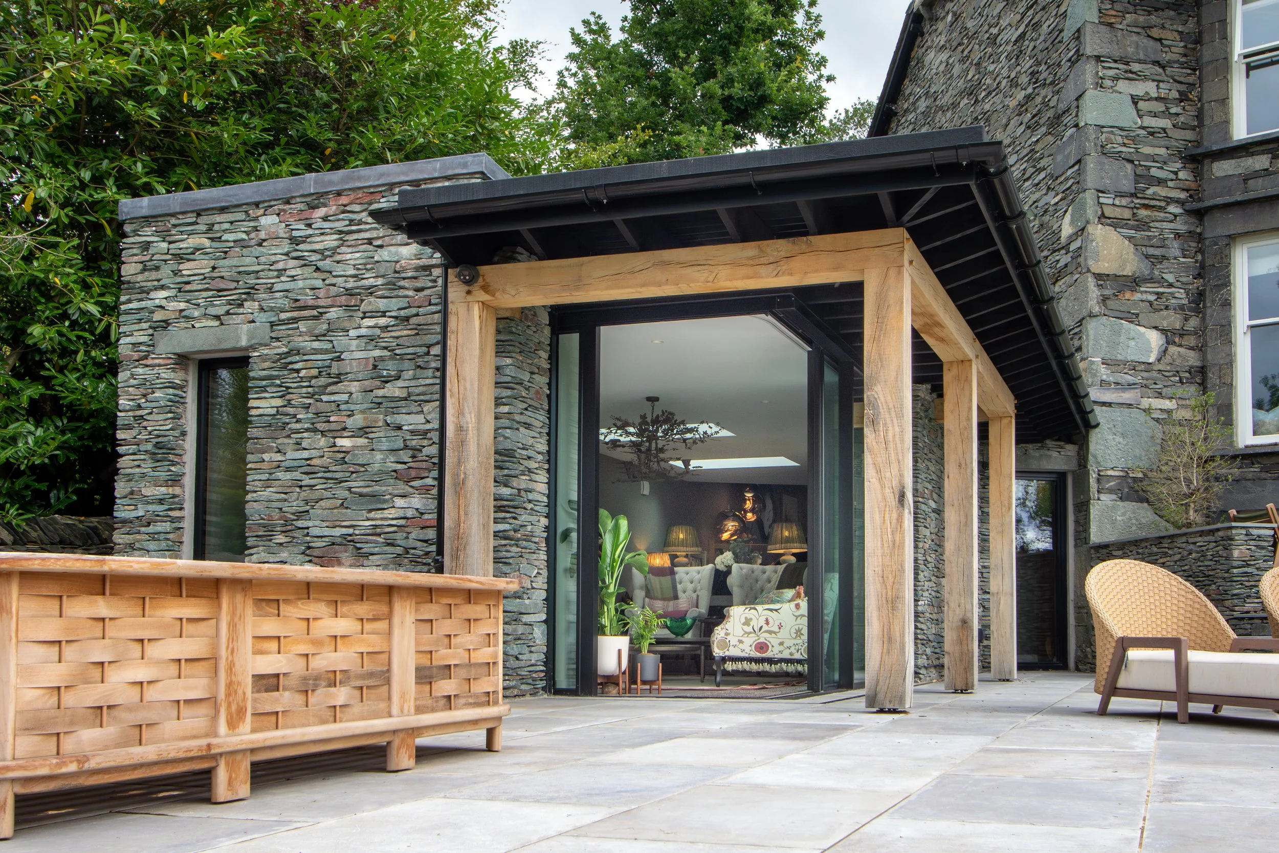 Exterior view of a modern house with stone walls, large glass doors, and wooden support beams, featuring a patio area with outdoor furniture and plants.