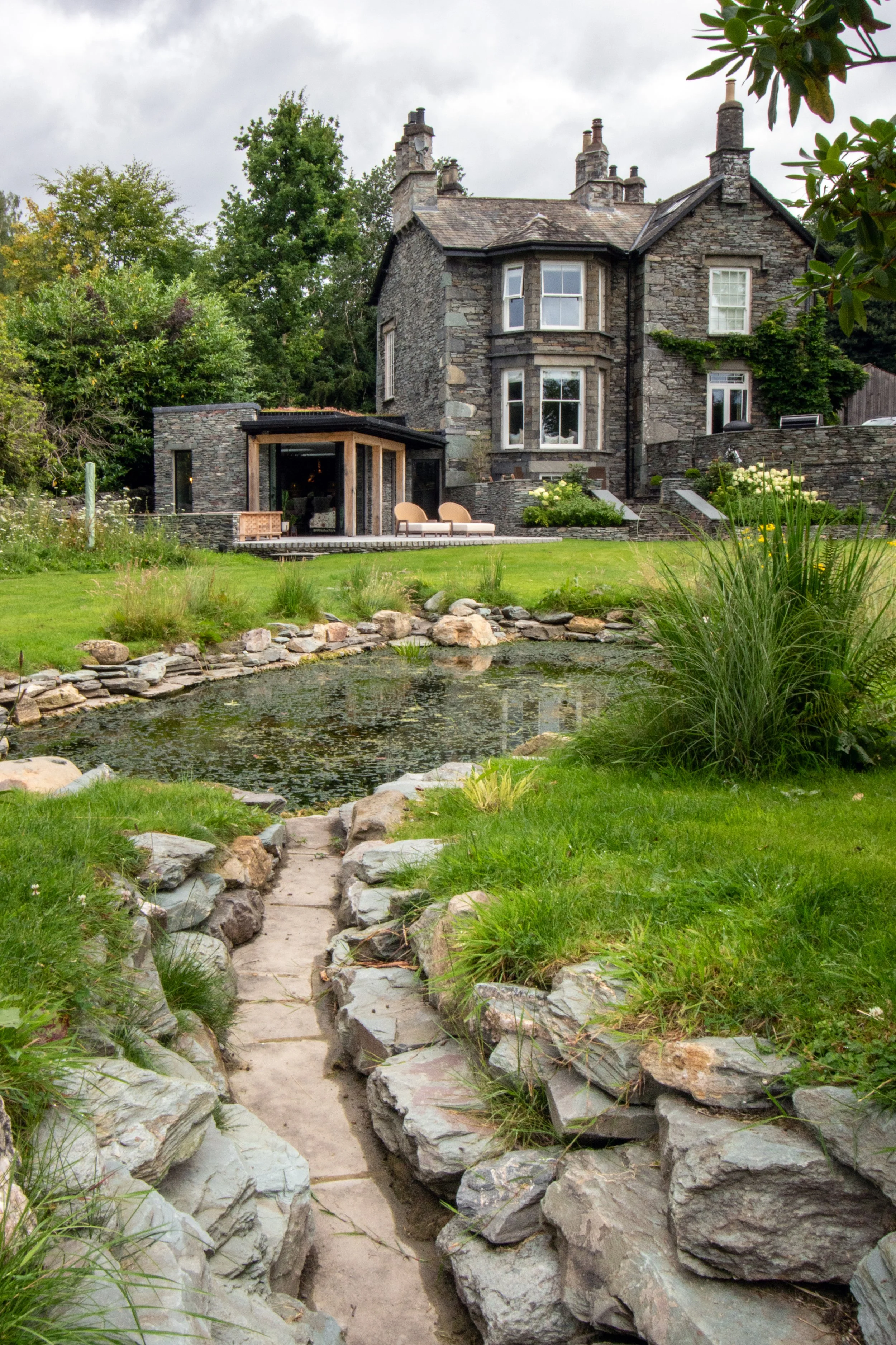 A stone pathway leading to a pond with rocks and greenery, with a large stone house and a modern glass extension in the background under a cloudy sky.