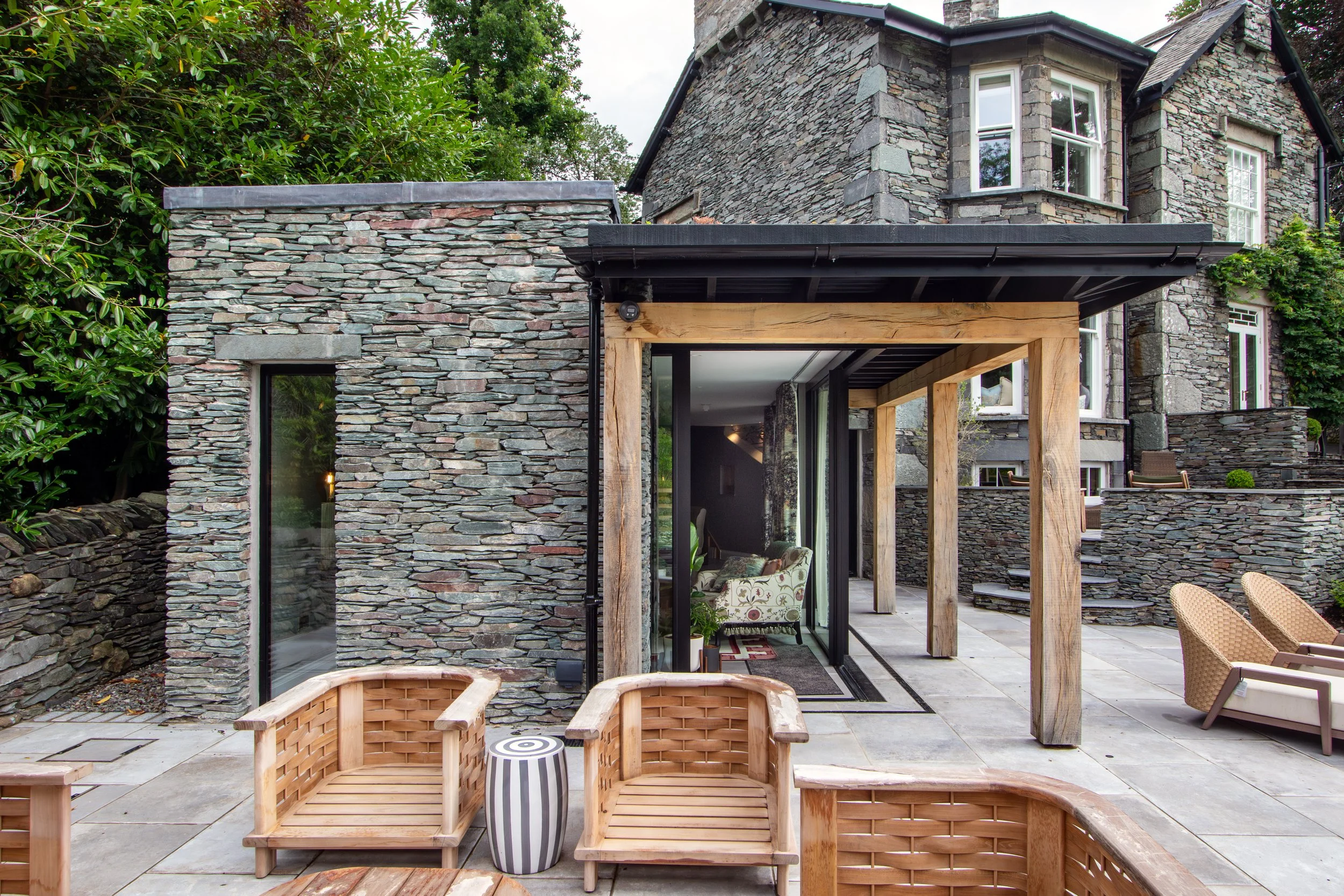 Modern stone house with a patio featuring wooden and wicker outdoor furniture, leading to a living room through glass sliding doors, surrounded by greenery.
