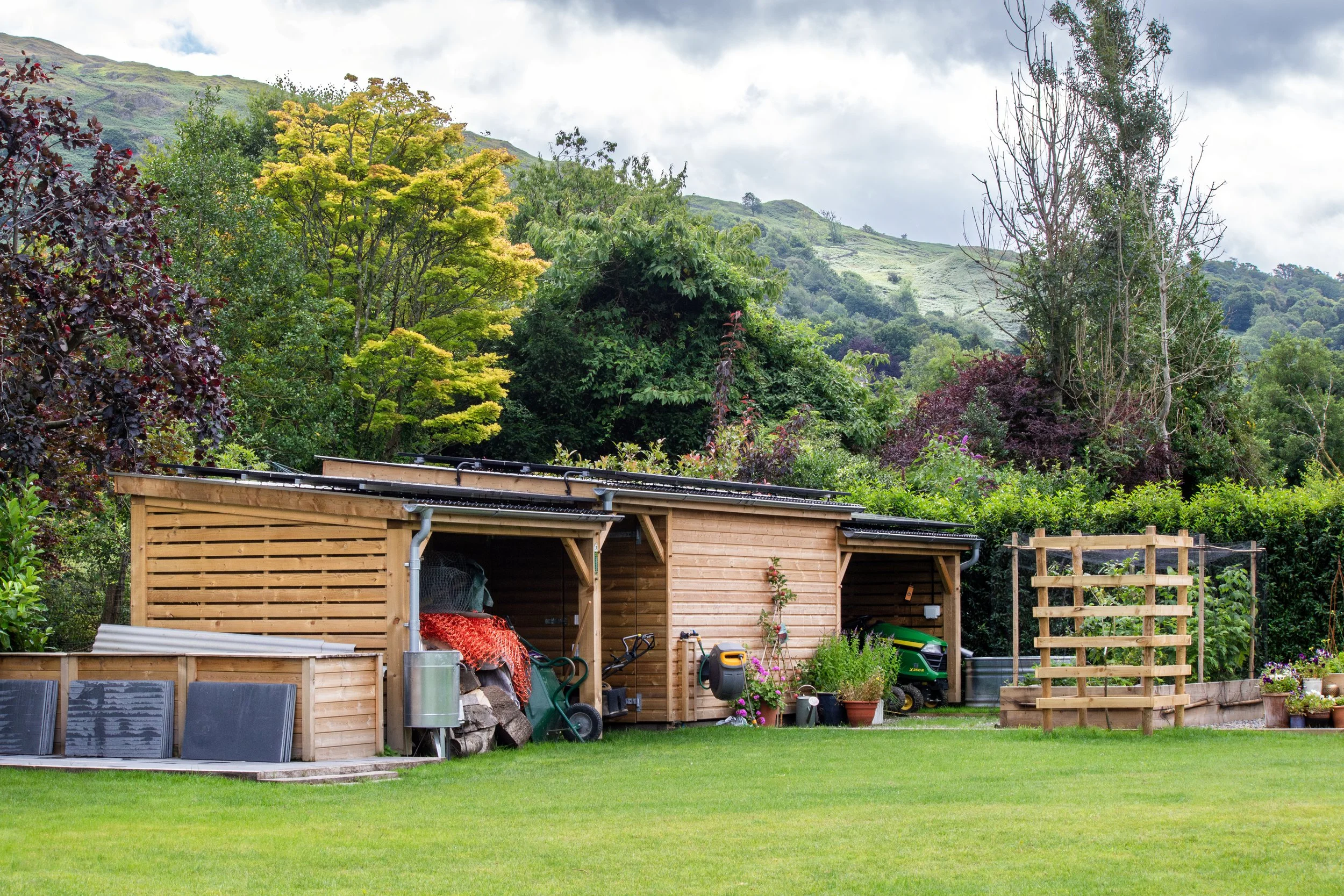 A garden shed made of wood, positioned on a well-maintained lawn with trees and hills in the background. The shed contains gardening tools, plants, and a small tractor.