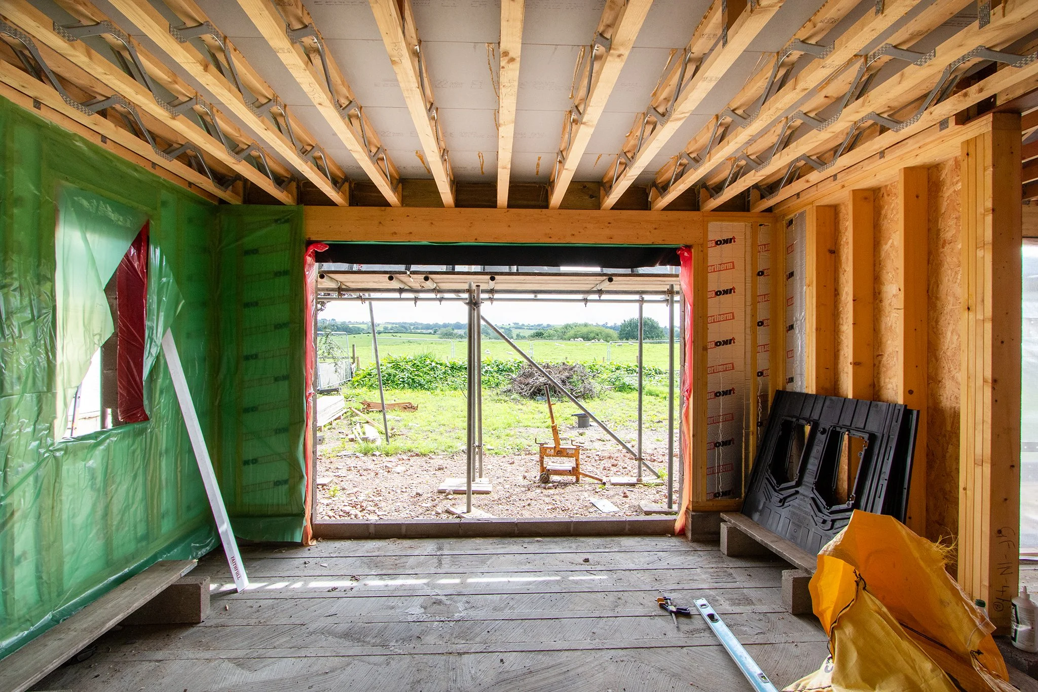 Interior view of a house under construction with wood framing, open view of the outdoors, construction tools on the floor, and construction materials leaning against the walls.