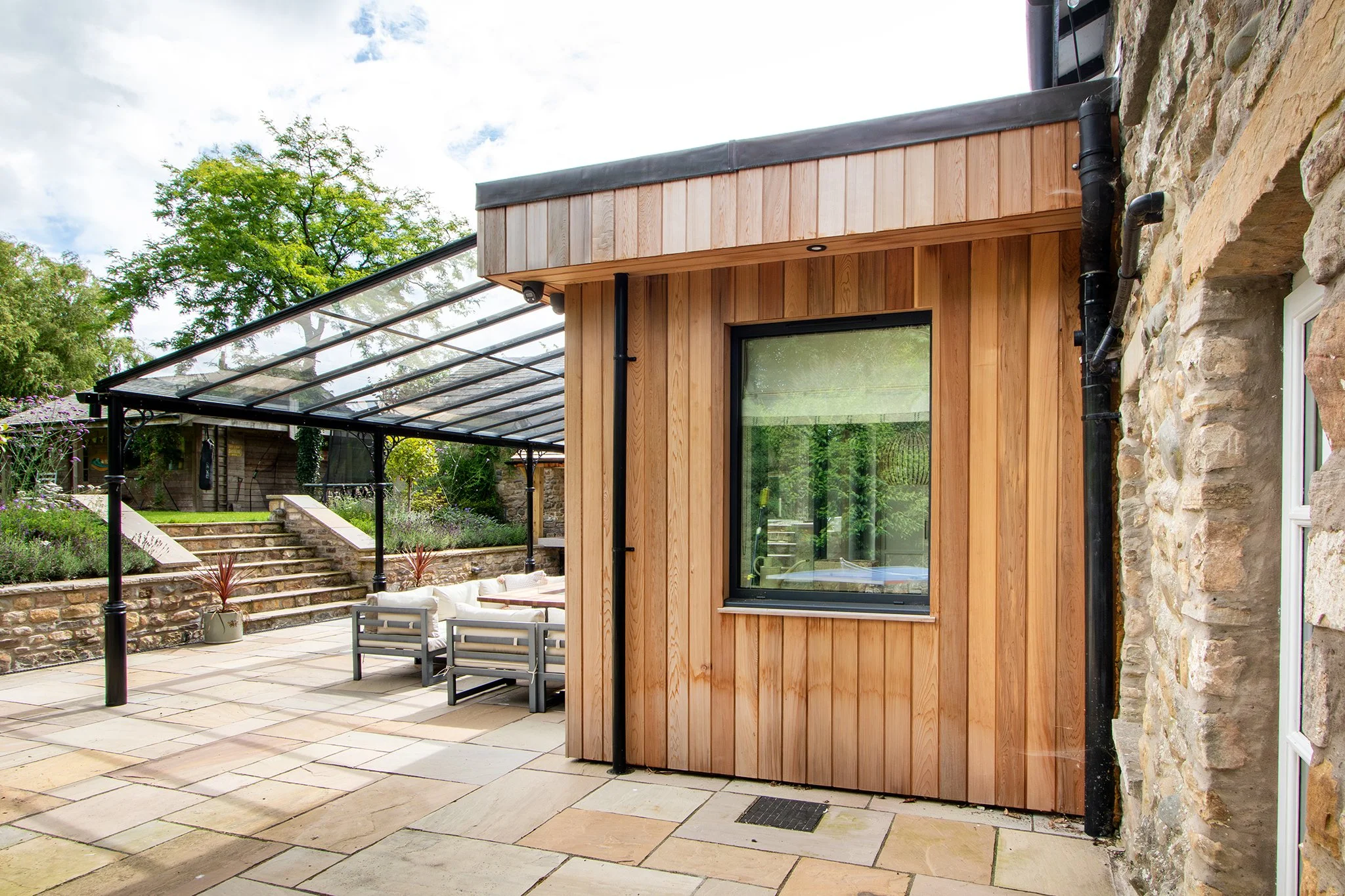 Modern outdoor patio area with stone paving, a glass-roofed canopy, and a small wooden structure with a window, surrounded by greenery and stone stairs.