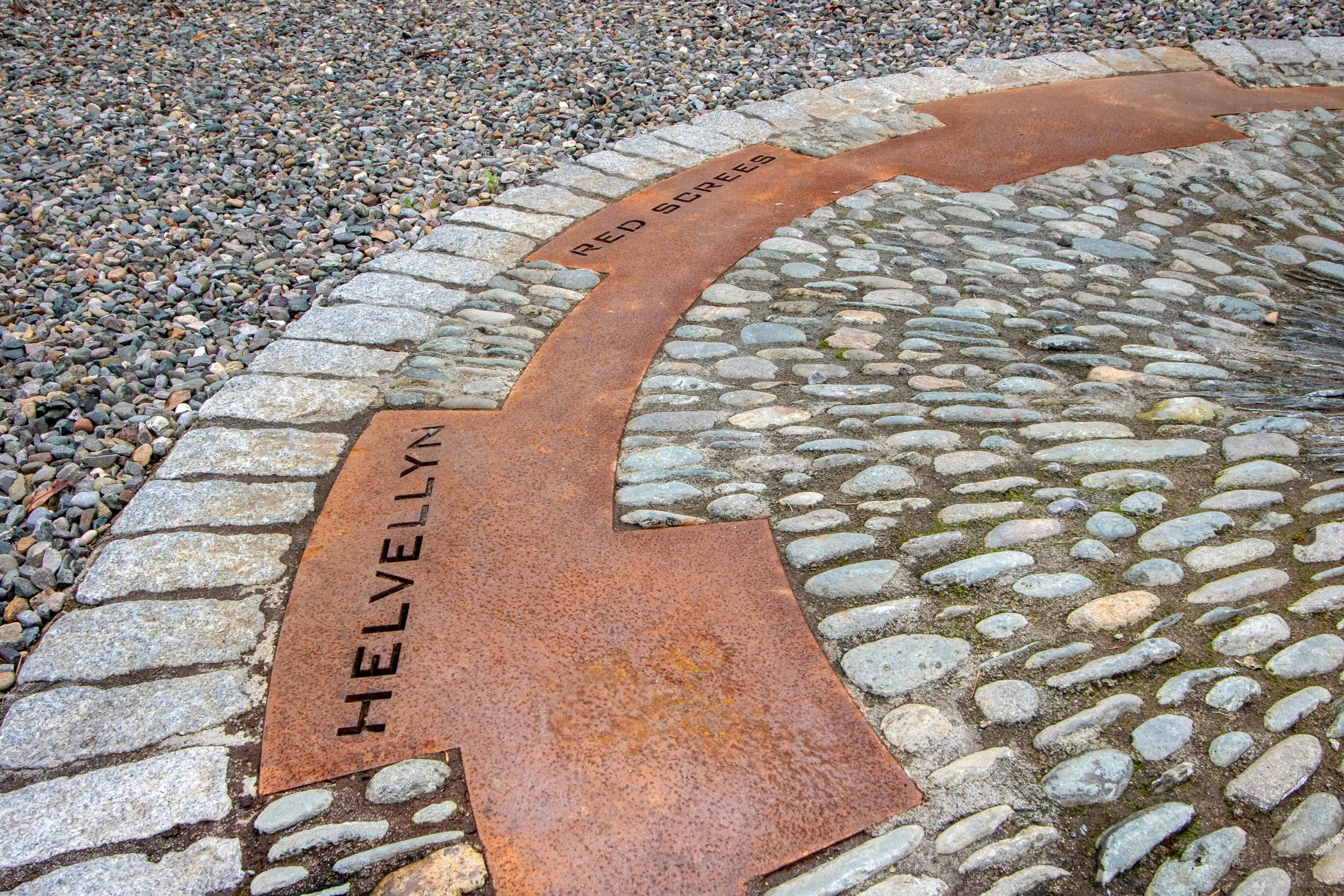 A curved metal plaque embedded in cobblestone pavement, labeled 'HELLEVLYN RED SQUARE' with surrounding stones and gravel.