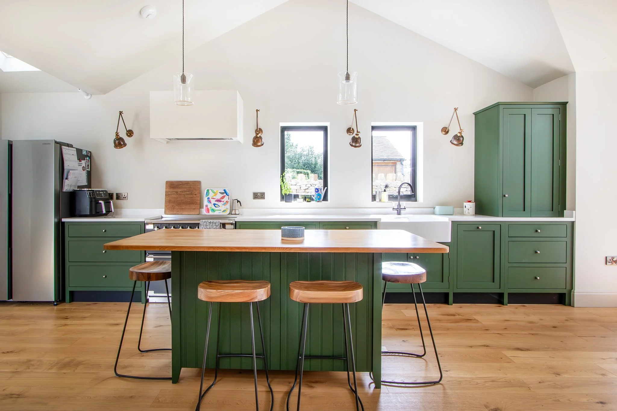 Modern kitchen with green cabinets, wooden island, three bar stools, stainless steel refrigerator, and two windows.