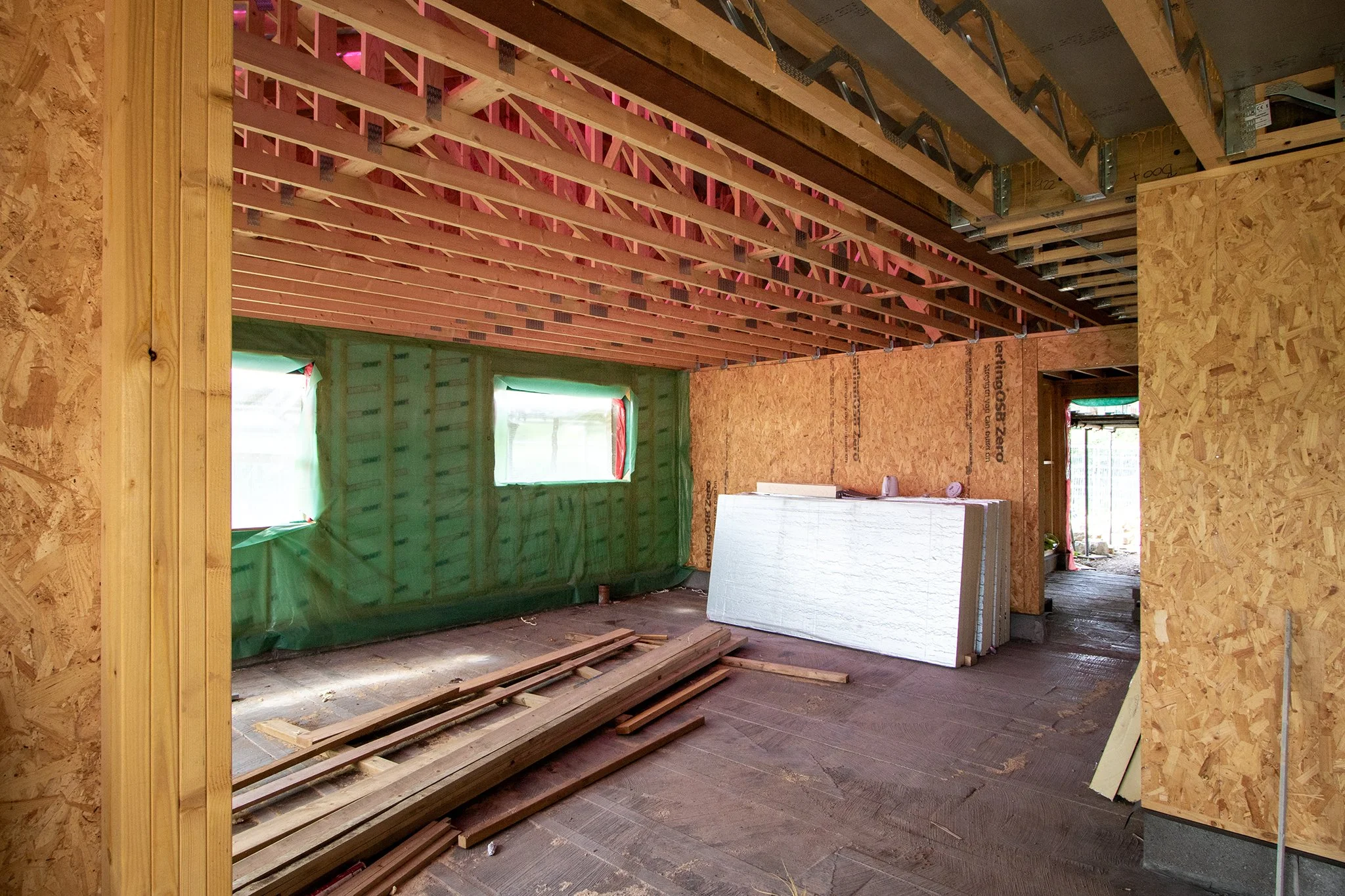 Interior view of a house under construction, showing exposed wooden framing, green plastic-covered windows, and construction materials like insulation panels and lumber on the floor.