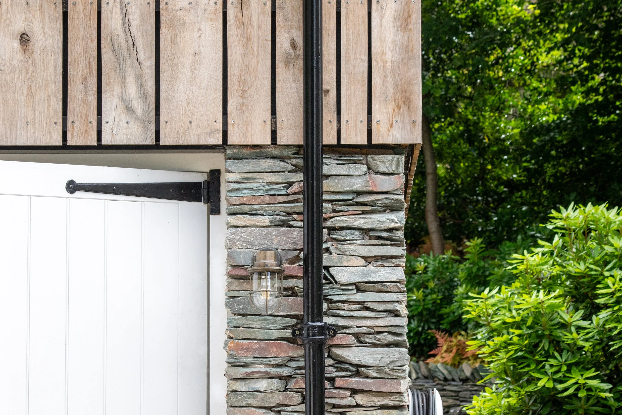Close-up of a modern house corner with a white garage door, stone wall, black vertical pipe, and outdoor light fixture, surrounded by green foliage.
