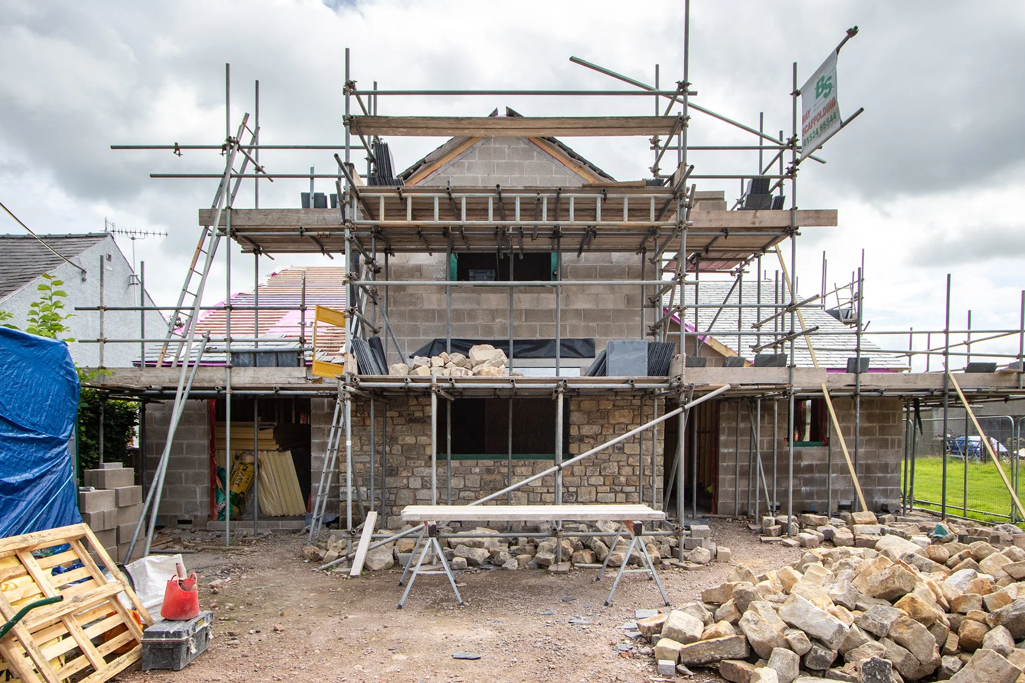 A two-story house under construction with scaffolding surrounding it, construction materials and rocks scattered on the ground, and some partially finished walls.