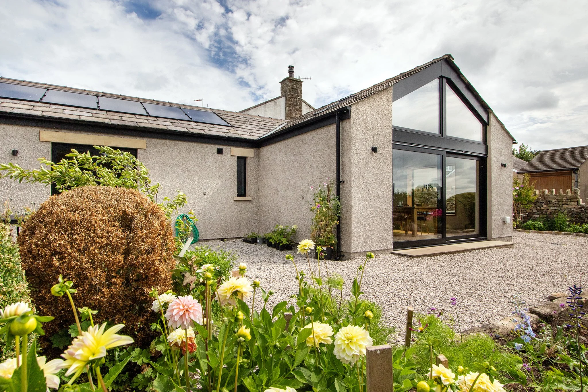 A modern house with a large glass sliding door and solar panels on the roof, surrounded by a garden with blooming flowers.