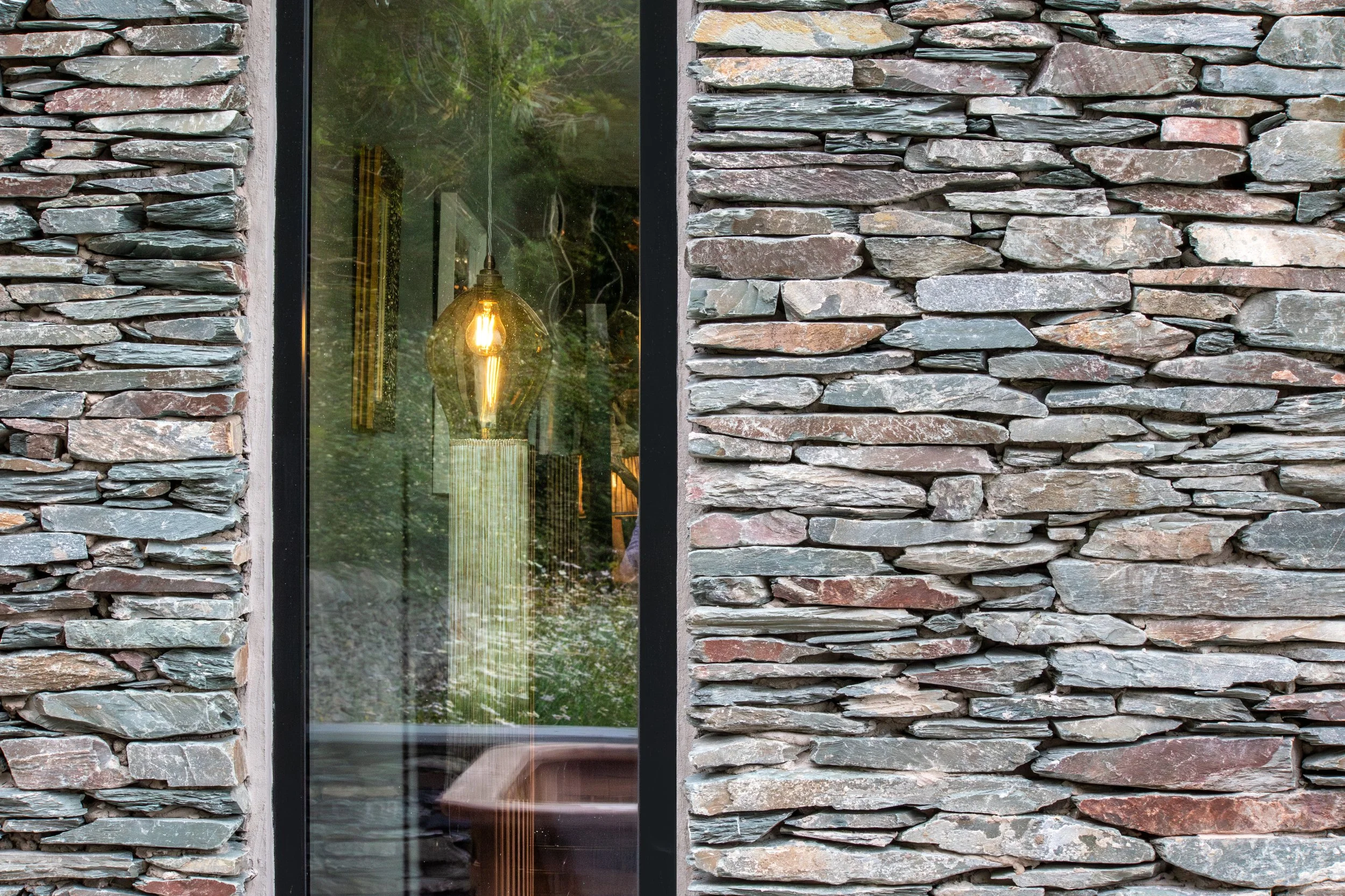 View of a stone wall with a large glass window, reflecting greenery outside and showing an interior light fixture inside.