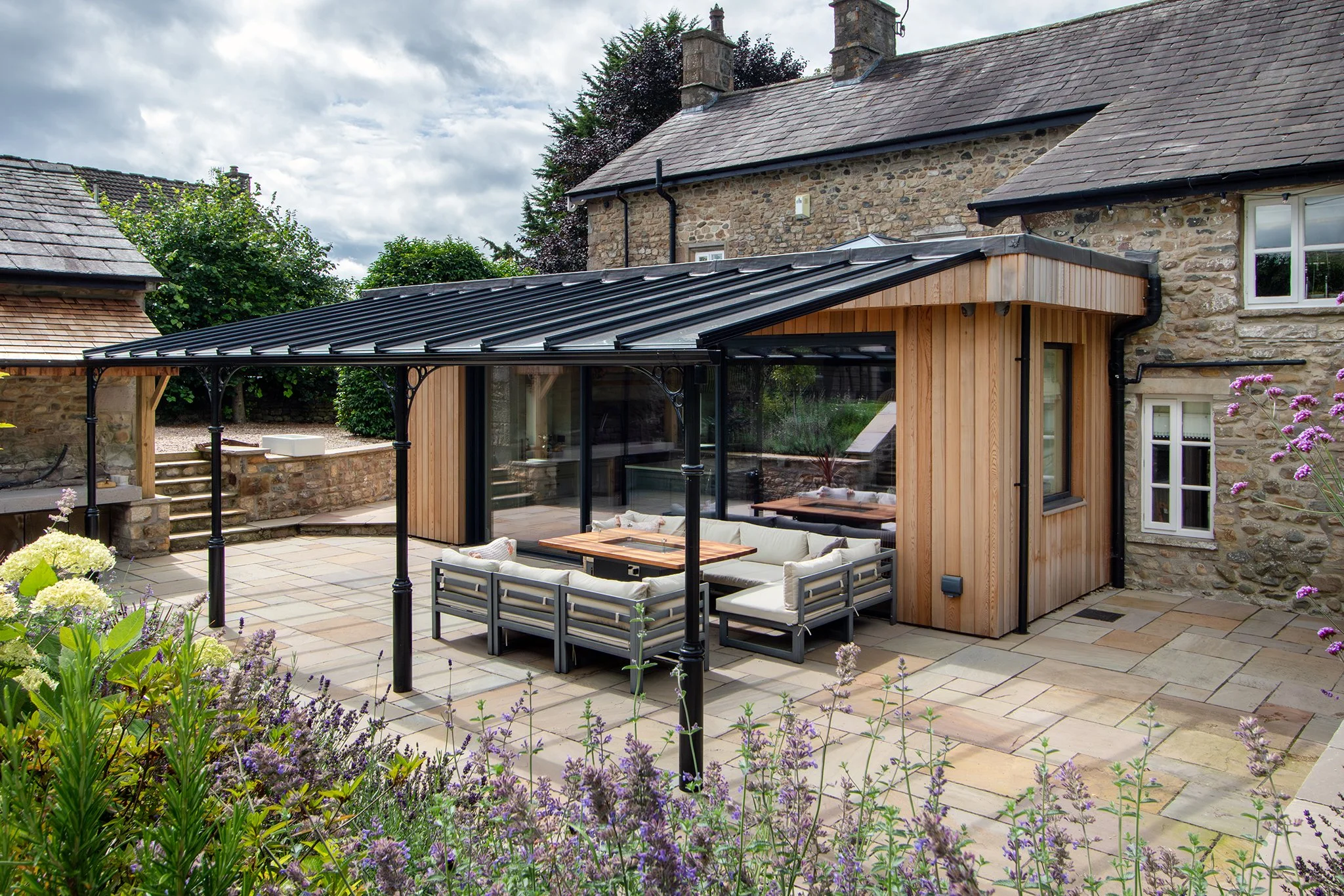 A modern outdoor patio with a glass-enclosed seating area, stone and wood exterior, and flowering plants in the foreground.