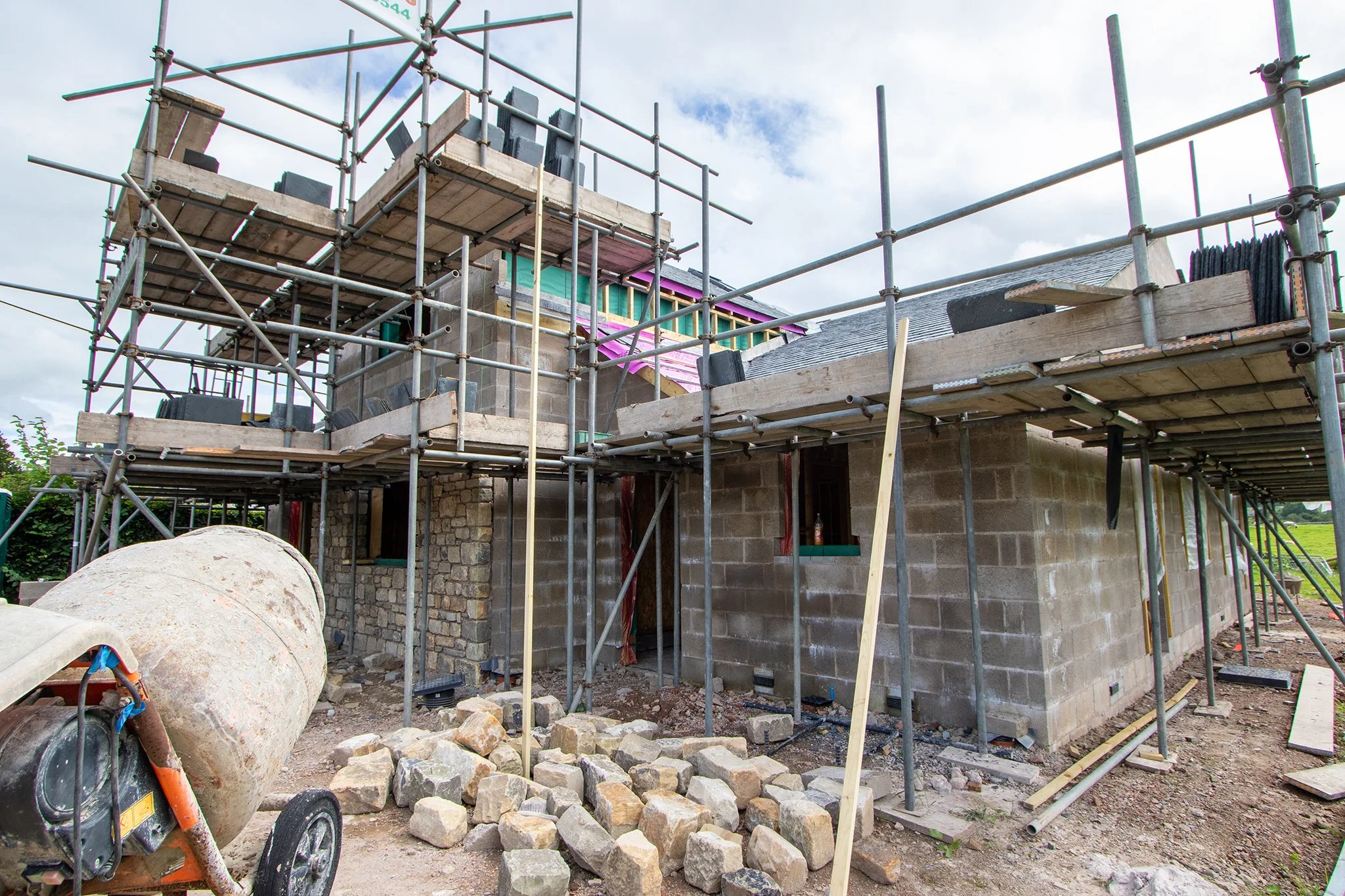 Construction site with scaffolding surrounding a partially built brick house, rocks and construction materials on the ground, and a cloudy sky.