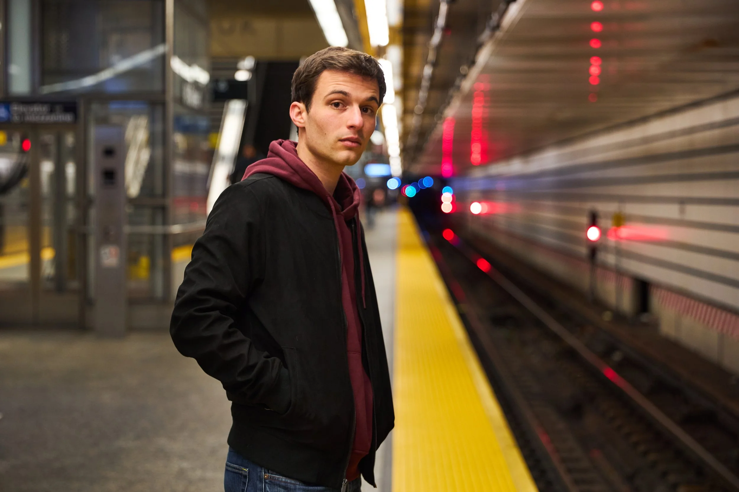 Young man standing on a subway platform at night, facing the camera with hand in pocket, with train tracks and blurred train in the background.