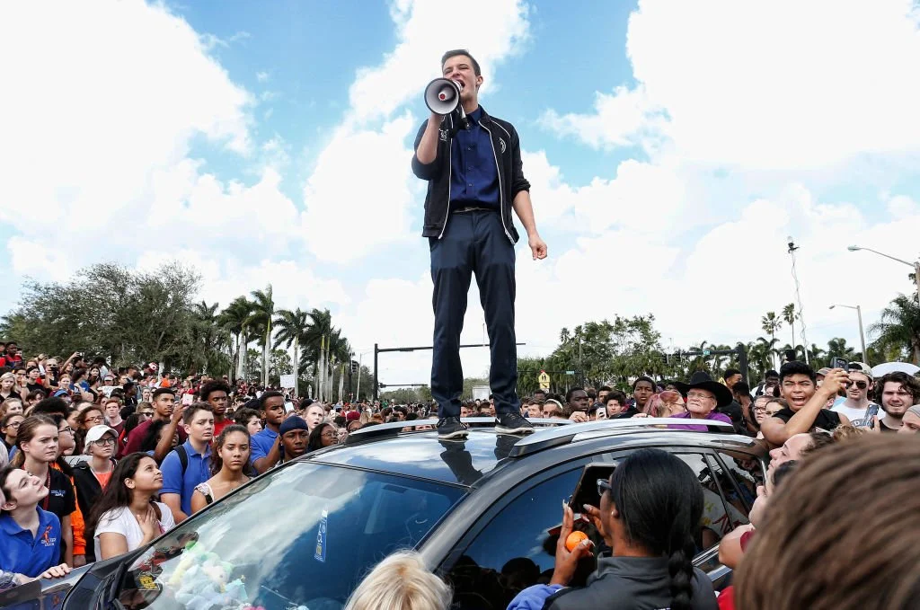 A man standing on a car roof with a megaphone addressing a large crowd of people outdoors under a cloudy sky.