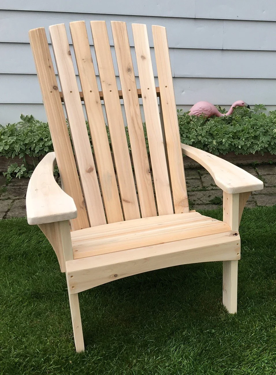 A wooden Adirondack chair placed on green grass in a garden with a small pink flamingo garden ornament and green foliage in the background.
