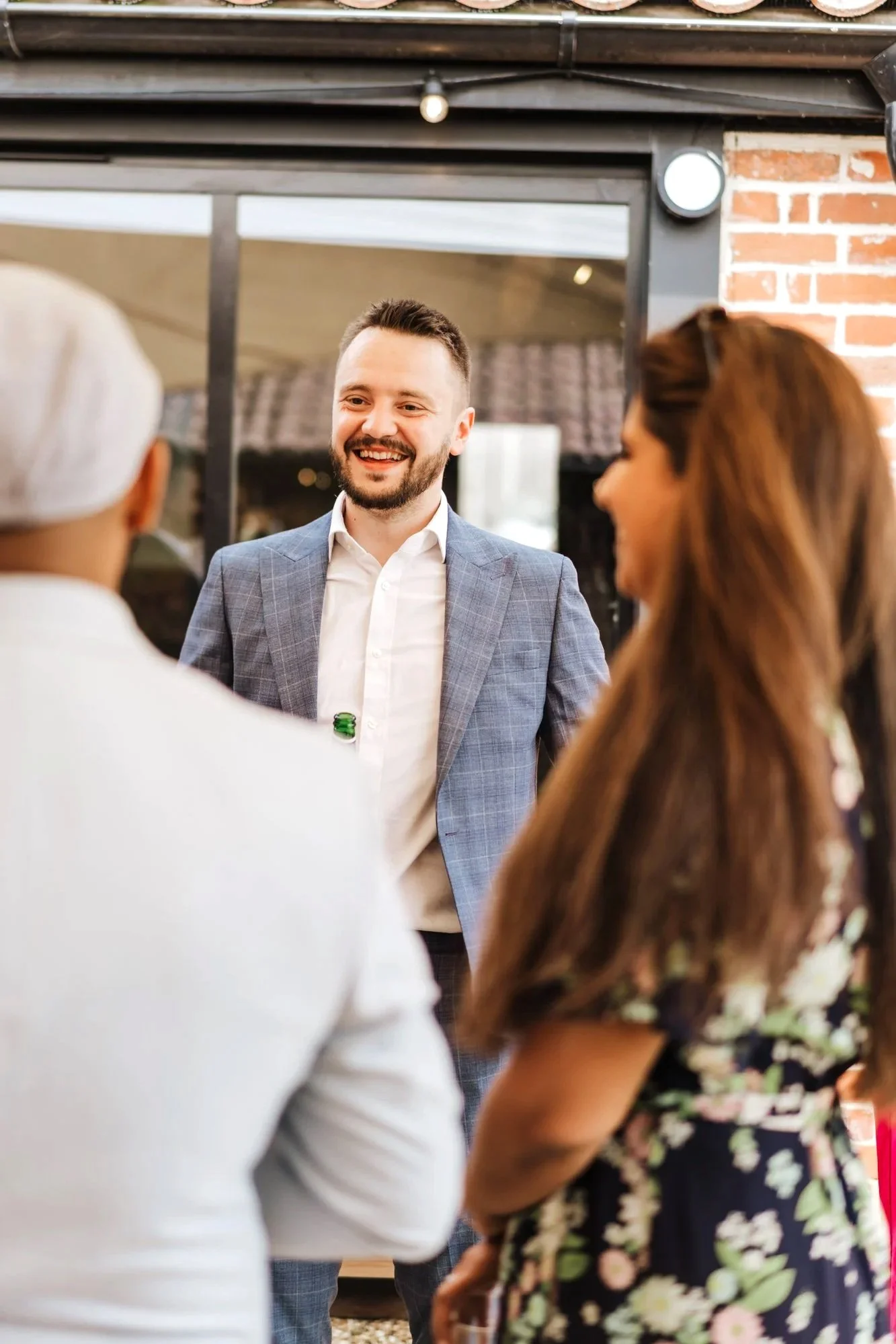 Three people engaged in conversation at an indoor event, with a man in a gray blazer smiling at two women, one in a white outfit and the other in a floral dress.