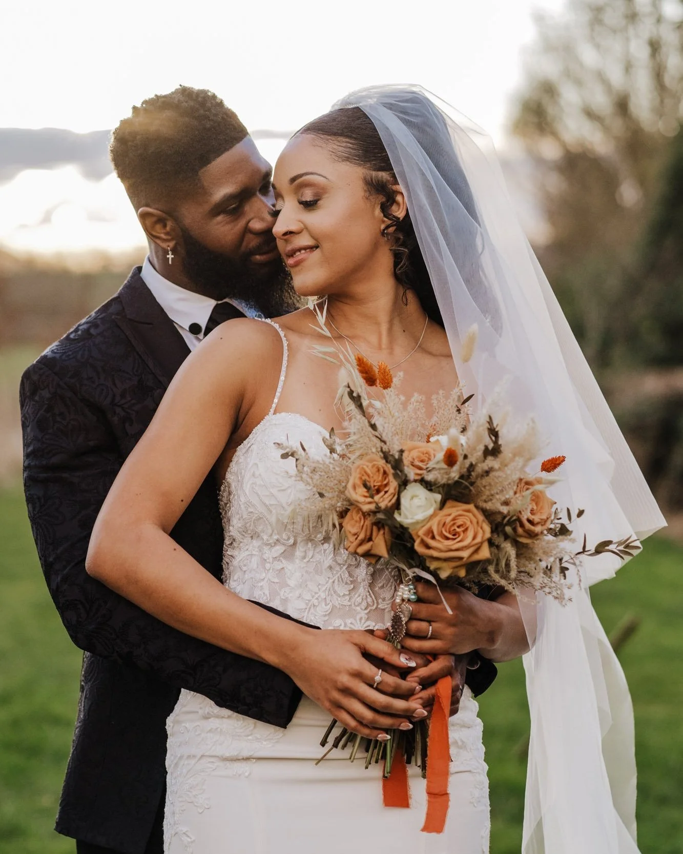 A newlywed couple standing outdoors, embracing and holding a bouquet of flowers, with the bride wearing a white lace wedding dress and veil, and the groom dressed in a black suit, during sunset.