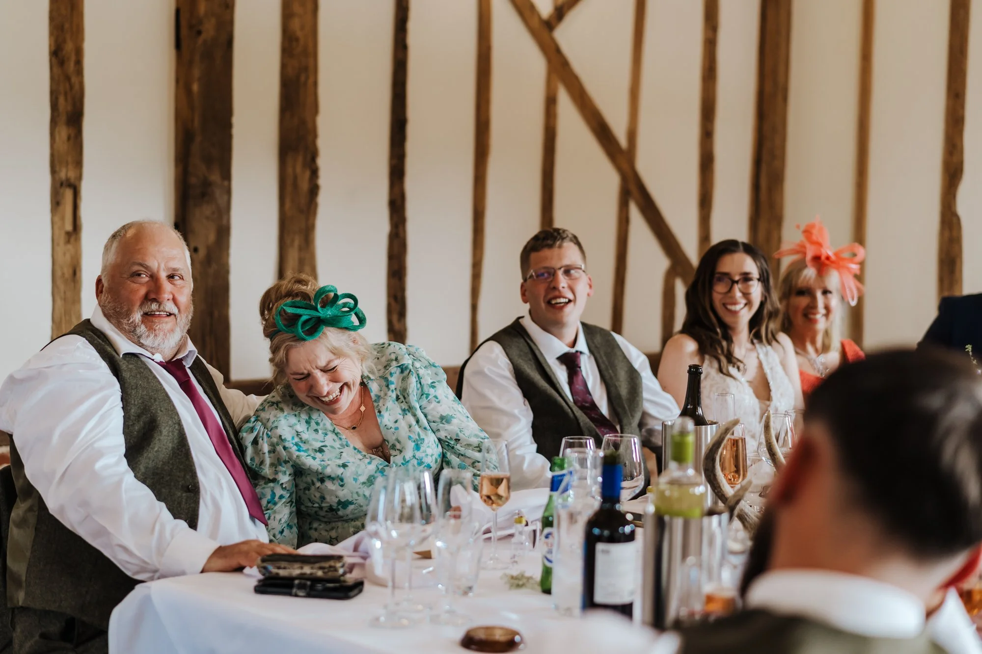 People sitting at a long dining table during a celebration, laughing and smiling, with rustic wooden beams in the background.