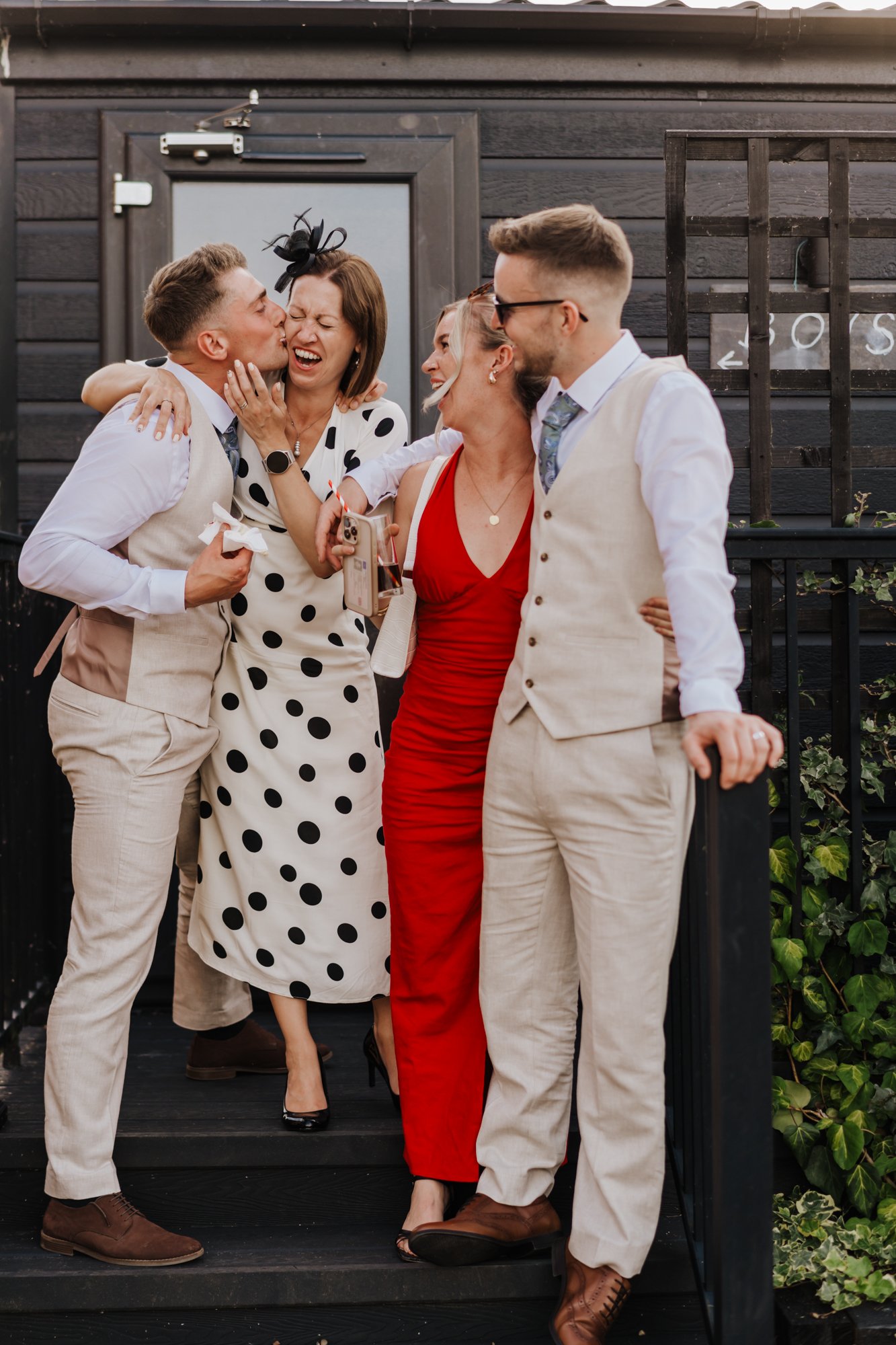 A group of five adults, three men and two women, celebrating outdoors in formal attire, with some hugging, laughing, and one kissing a woman on the cheek.