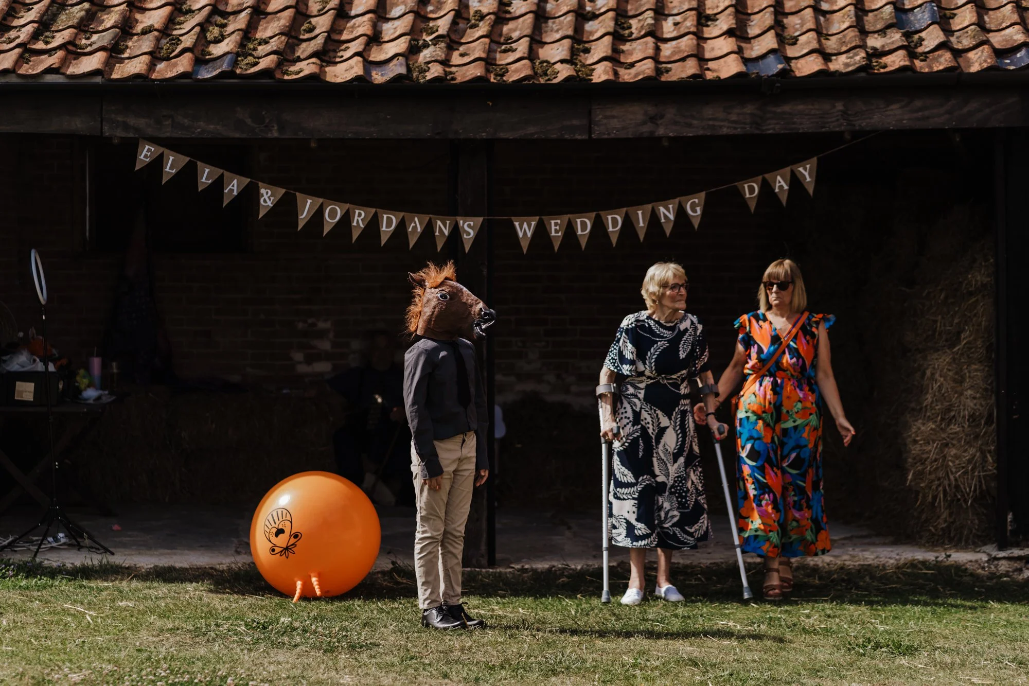 People attending a wedding celebration outdoors, including a person in a horse mask, and two women with walkers, standing in front of a barn with a banner that reads 'Ella & Jordan's Wedding Day.' There is also a large orange balloon decorated with a butterfly.