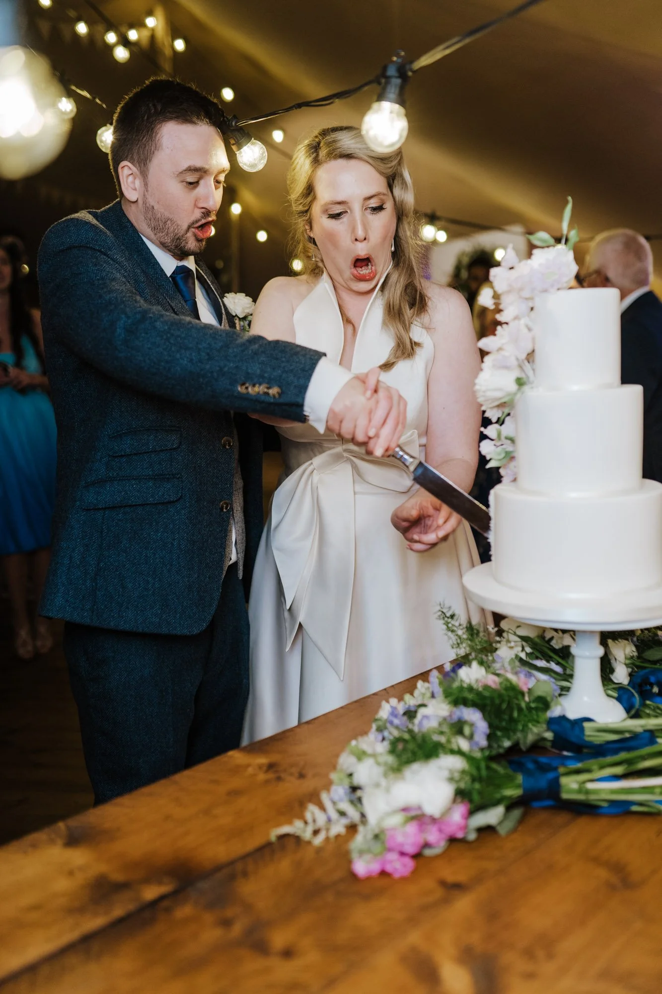 A bride and groom cutting a wedding cake together during their wedding reception, surrounded by floral decorations and string lights.