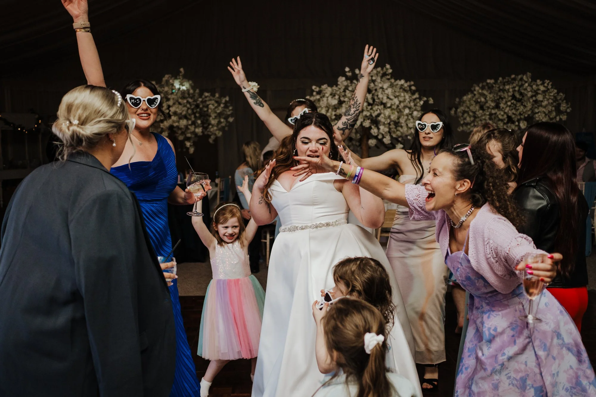 People celebrating at a wedding reception, with the bride in a white dress surrounded by women and children, some wearing sunglasses and holding drinks, dancing and smiling.