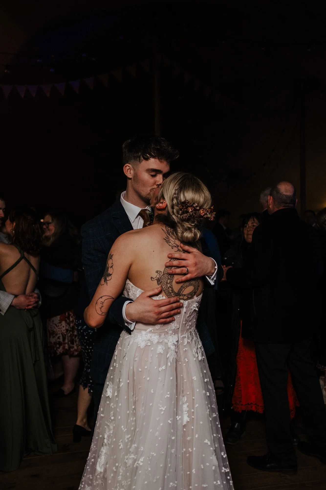 A couple dancing closely at a wedding reception, with the man in a dark plaid suit and the woman in a white lace wedding dress with tattoos on her back and arms, surrounded by guests in a dimly lit venue.
