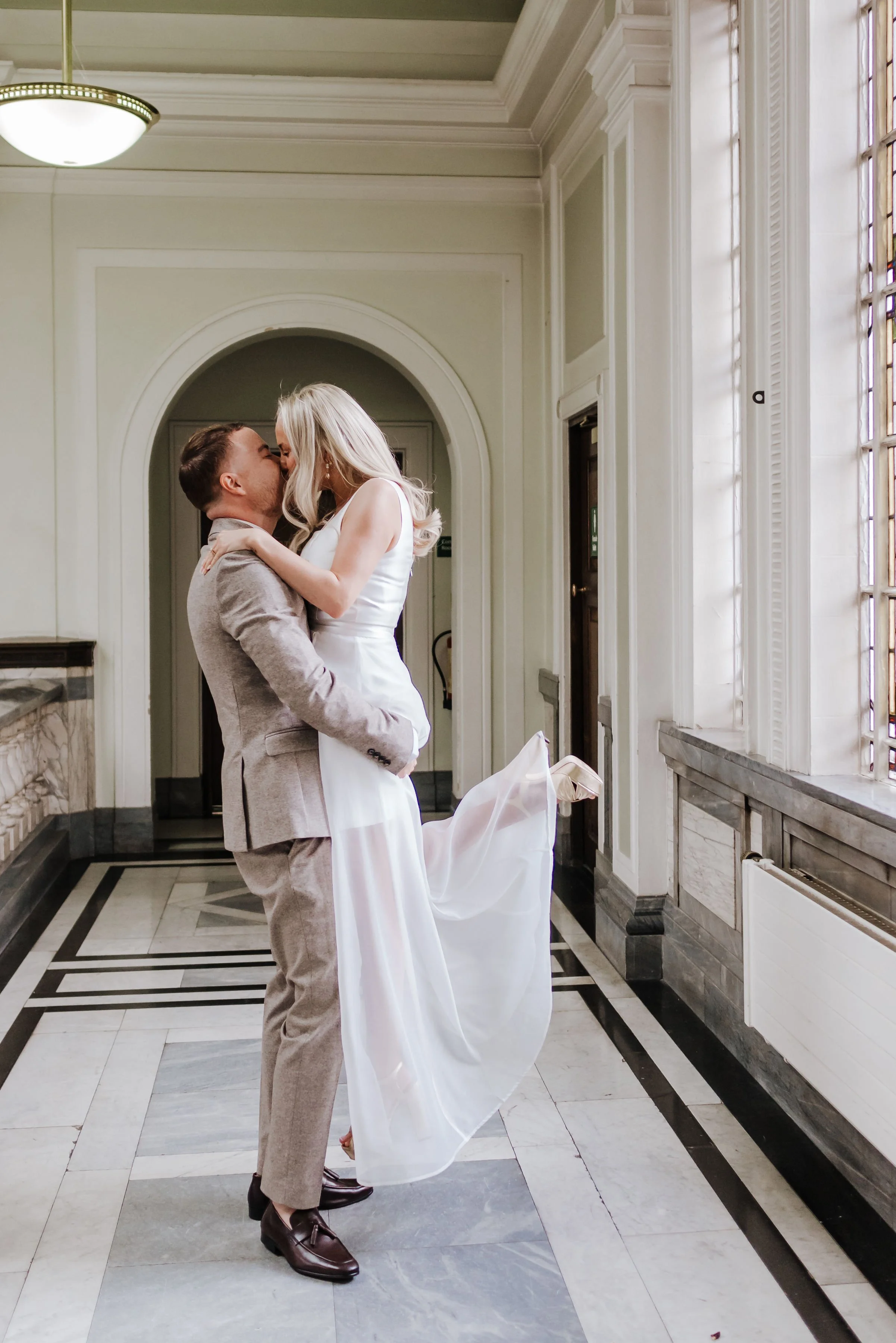 A couple dressed in wedding attire sharing a kiss inside a grand, vintage-style building with large windows and ornate architectural details.
