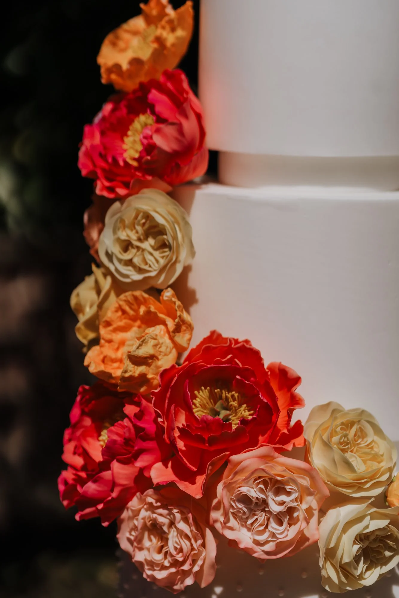 Close-up of a white cake decorated with a colorful arrangement of peony and rose flowers in shades of red, pink, cream, and orange.