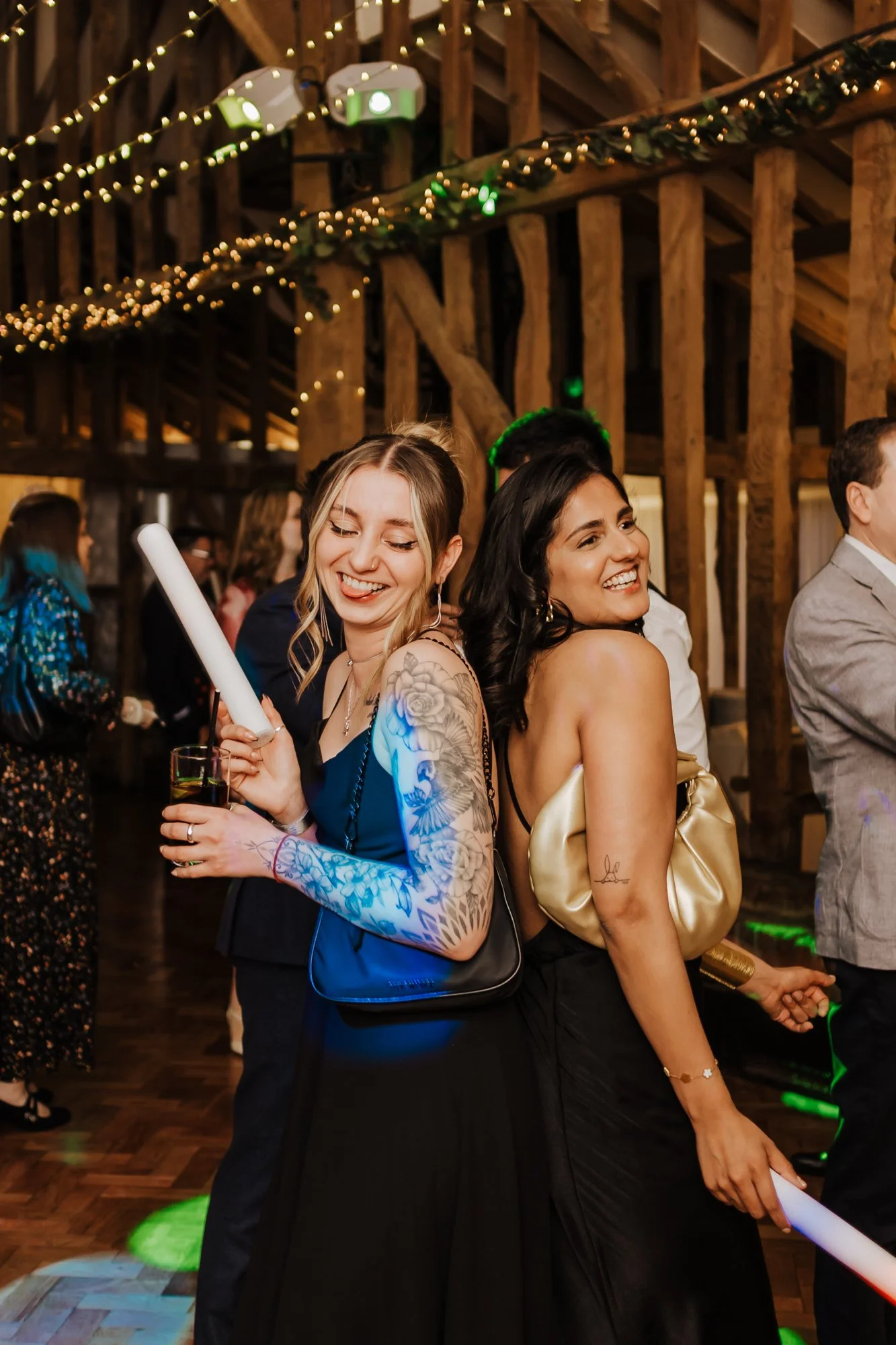 Two women smiling back-to-back at a party with string lights and wooden beams in the background.
