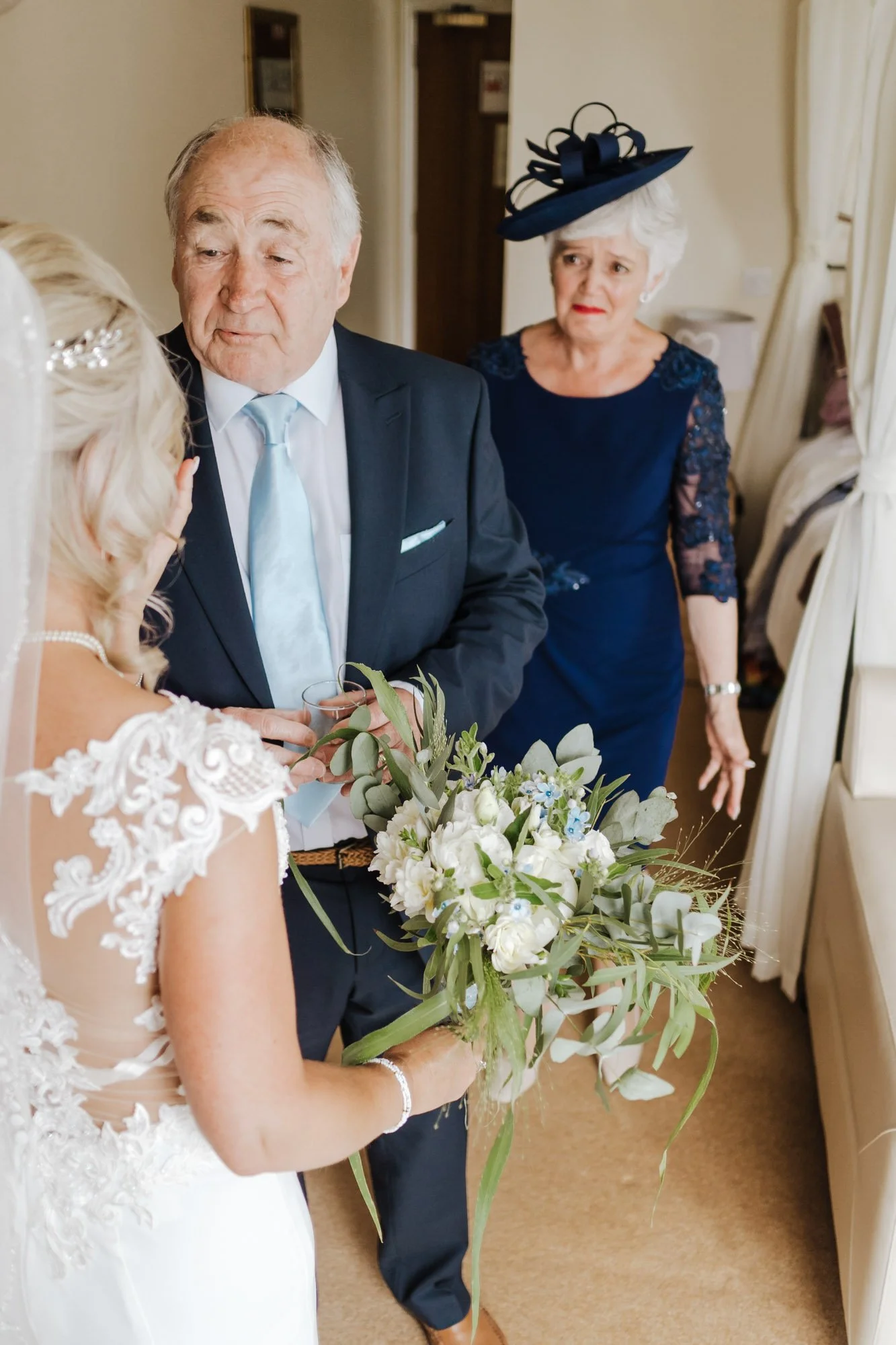 A bride holding a bouquet of white flowers and greenery, talking with an older man in a suit, with an older woman in a blue dress and hat standing behind them, in a room with beige walls and curtains.
