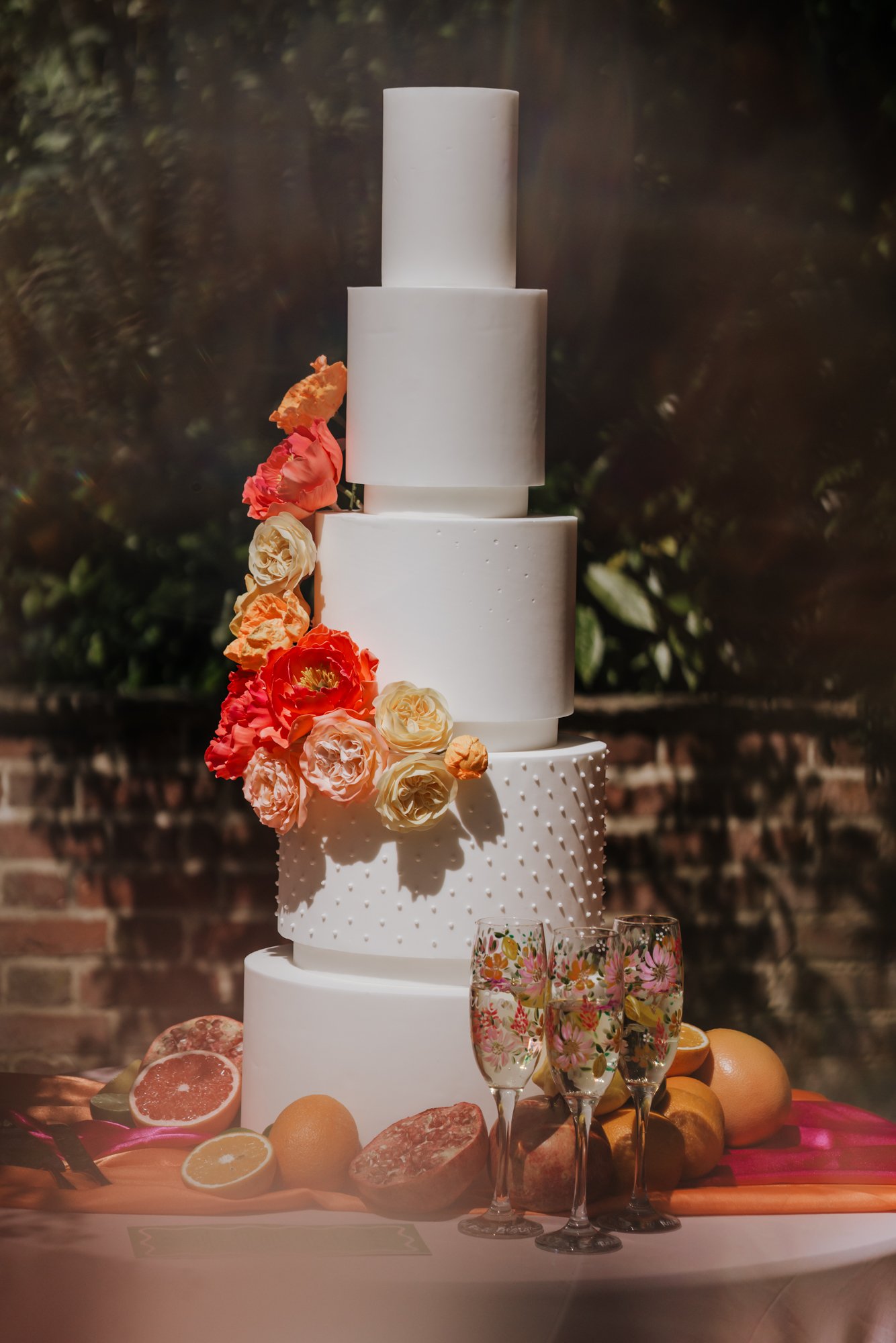 A multi-tiered white wedding cake decorated with fresh flowers and surrounded by citrus fruits and three champagne glasses.